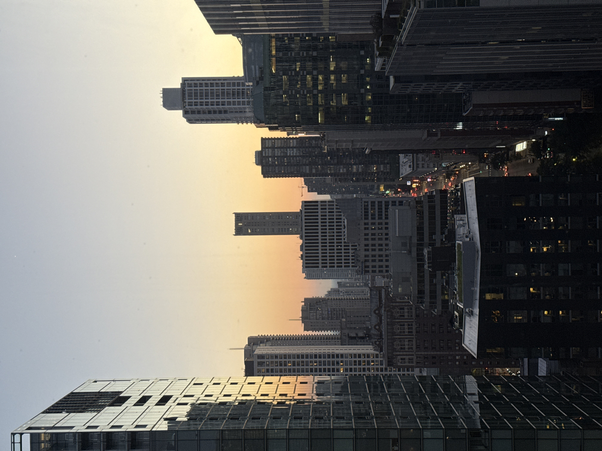 This is a vertical, eye-level, medium shot of a cityscape in Chicago, United States of America, at dusk. The image is dominated by tall buildings, showcasing a mix of modern glass and steel structures with older, more traditional architecture. The sky transitions from a pale, clear blue at the top to a warm orange and yellow gradient near the horizon, indicating the setting sun.
In the foreground on the left, a large, modern building with a glass facade reflects the warm tones of the sky. Its numerous windows are visible, and some appear to be lit from within. To the right of this, a dense collection of skyscrapers rises. Some have illuminated windows, suggesting activity within. The buildings vary in height and architectural style, creating a layered effect. A street is visible winding through the city in the mid-ground, with tiny car headlights and taillights hinting at ongoing traffic.
Further into the distance, more buildings continue to stretch towards the horizon, some appearing as silhouettes against the fading light. The overall impression is one of a bustling urban environment at the close of day, with the city lights beginning to twinkle as darkness approaches. The scene is devoid of any visible people or immediate action.