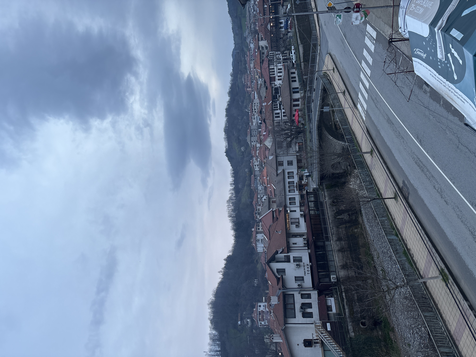 The image depicts a street scene in Zlatograd, Bulgaria. The view is angled downward from a high vantage point, showing a winding road with a pedestrian crosswalk. Along the right side of the road, there is a billboard advertising Davidoff Pulze. Opposite the road, houses are built along a hillside, their red roofs and white walls contrasting against the grey sky and the dark green of the hill. In the foreground, there is a railing along a walkway that runs parallel to the road, with bare tree branches visible. The sky is overcast with heavy clouds. The houses appear to be of traditional Balkan architectural style. Some signs in Cyrillic are visible on the buildings, one of which reads "ТОНИ" (TONI).