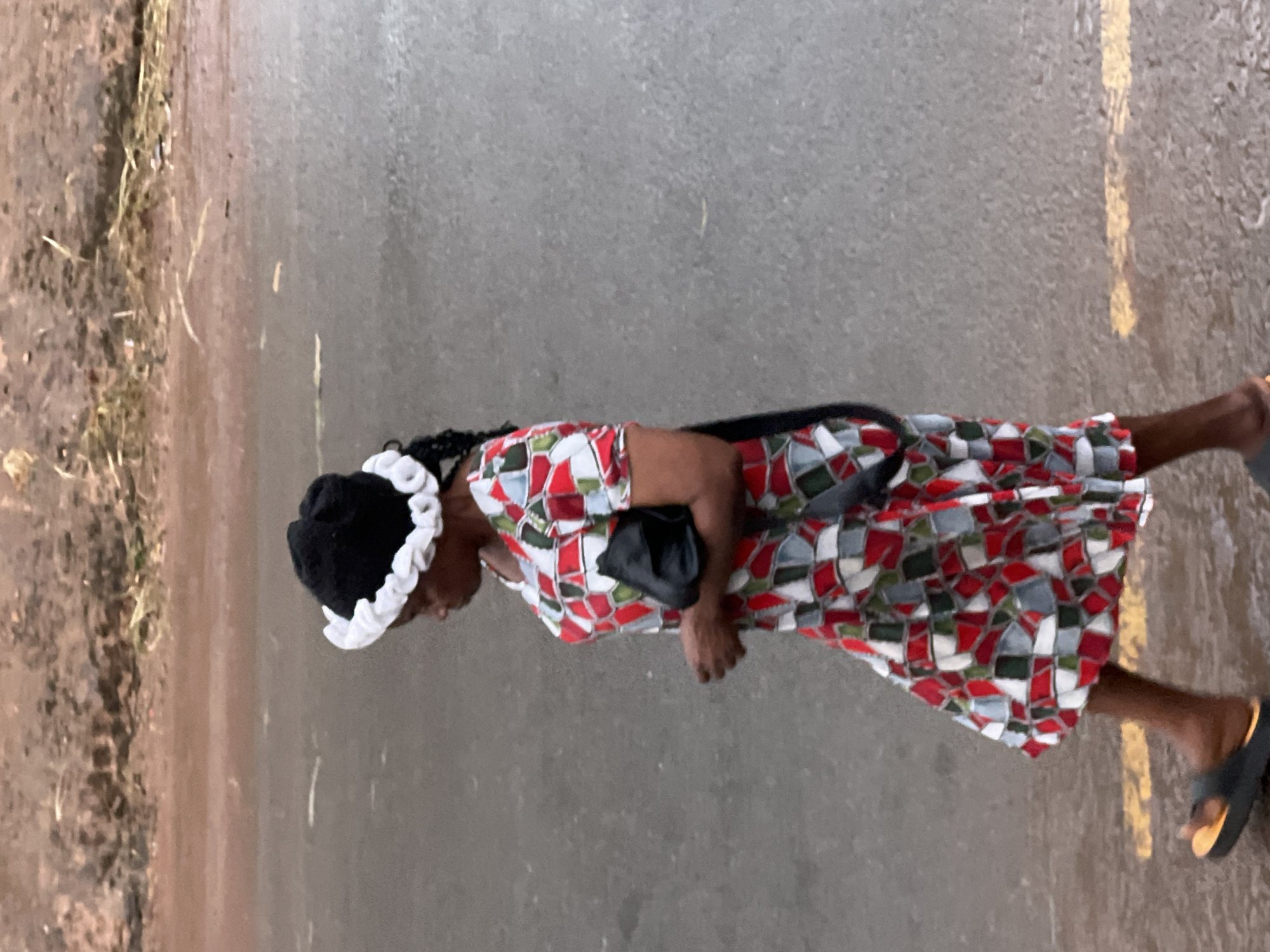 A woman wearing a black hat with a white trim and a colorful, geometric-patterned dress walks across a wet asphalt road in Nkpor, Nigeria. She carries a black clutch bag in her right hand. The road has yellow lines painted on it, and the background consists of a reddish-brown embankment with some dry vegetation. The lighting suggests it might be overcast or late in the day.
