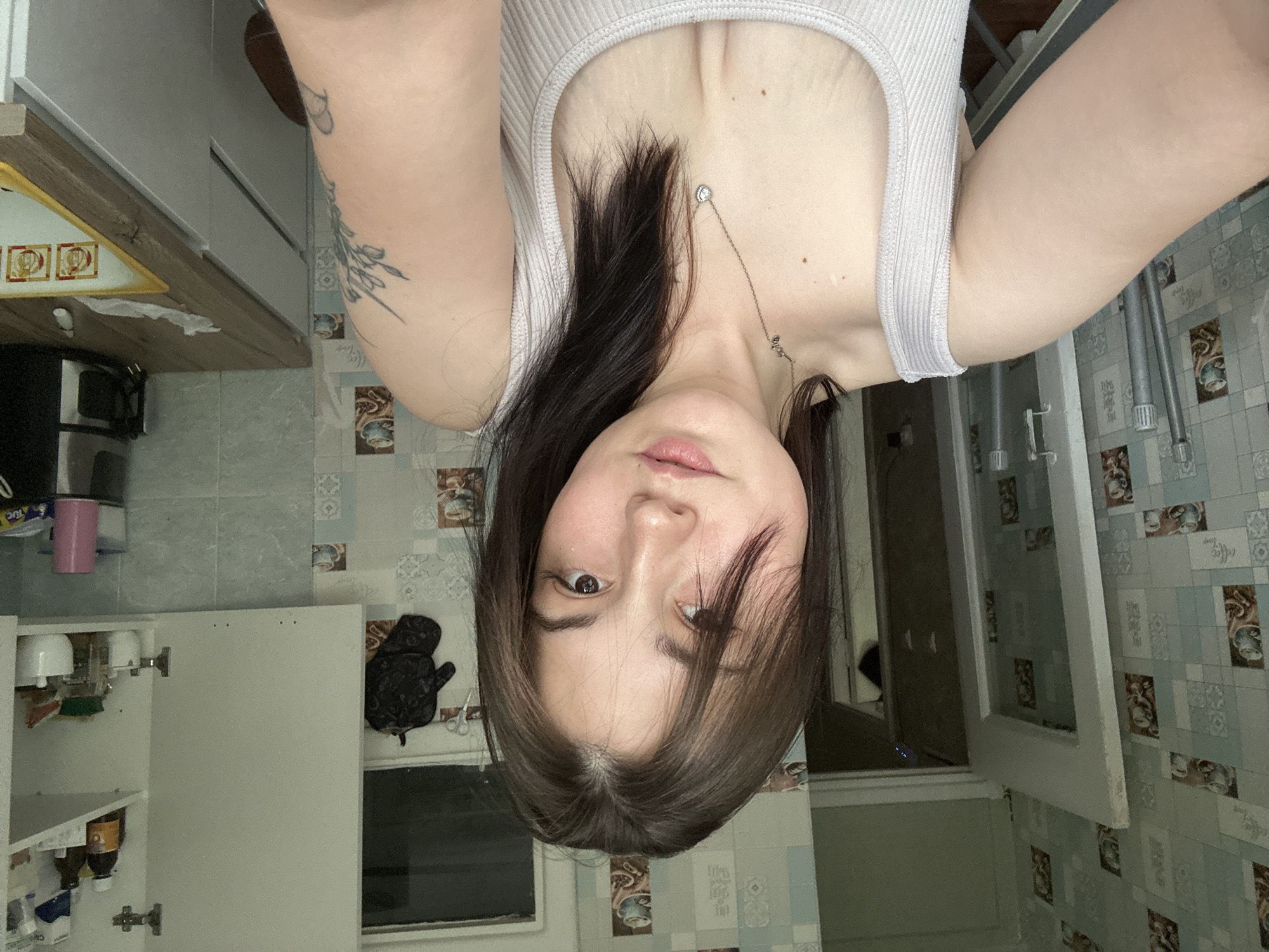 The image is a selfie taken by a young woman, oriented upside down. She is in what appears to be a bathroom or kitchen area, with tiled walls featuring a blue and white patterned design. In the foreground, a white cabinet with its door open reveals various bottles and containers inside. To the left, a portion of a countertop with a wooden edge is visible, and on it, a yellow object with three framed pictures on it. A black, soft object, possibly a stuffed toy, sits on a shelf next to the open cabinet.
The woman is wearing a light-colored ribbed tank top and a delicate necklace with a small pendant. She has long, dark hair that falls around her face. Her expression is neutral. On her left arm, a tattoo of floral or plant-like imagery is visible. The lighting suggests it could be daytime, but without clear windows or external views, the specific time of day is uncertain. The overall scene suggests an indoor, domestic setting. There is no visible text in the image.