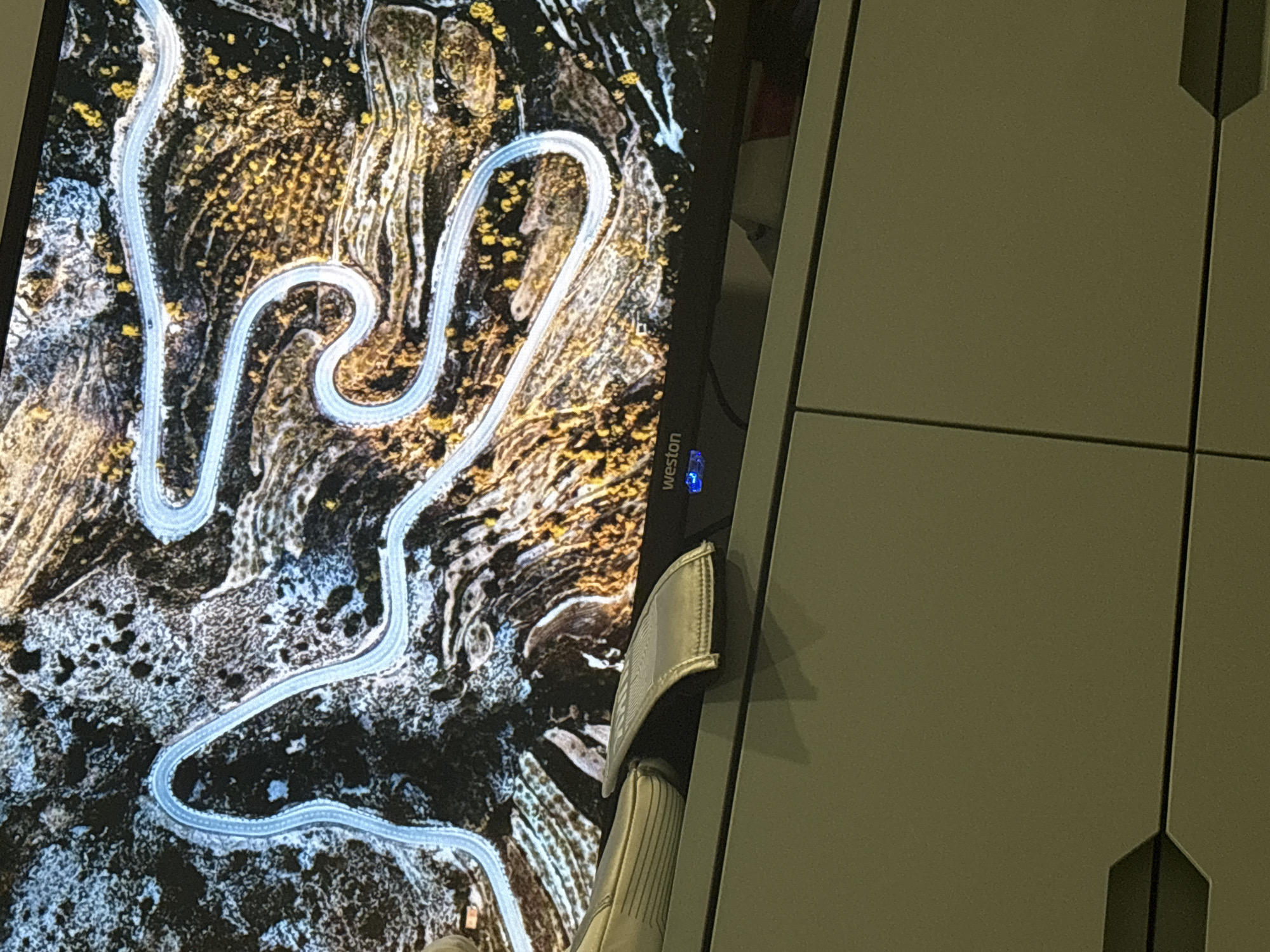 The image shows a portion of a monitor screen displaying an aerial view of a winding road through a mountainous terrain. The road is light-colored and contrasts with the dark and textured landscape. The landscape consists of varying shades of brown, black, and white, suggesting a mix of vegetation, rocks, and possibly snow. The monitor has a brand name "Weston" printed on its bezel. A partial view of a white and gray sandal is visible in front of the monitor. The background of the image consists of what appears to be a light-colored cabinet with vertical paneling. This does not appear to contain enough information to definitively state it is in Arinj, Armenia.