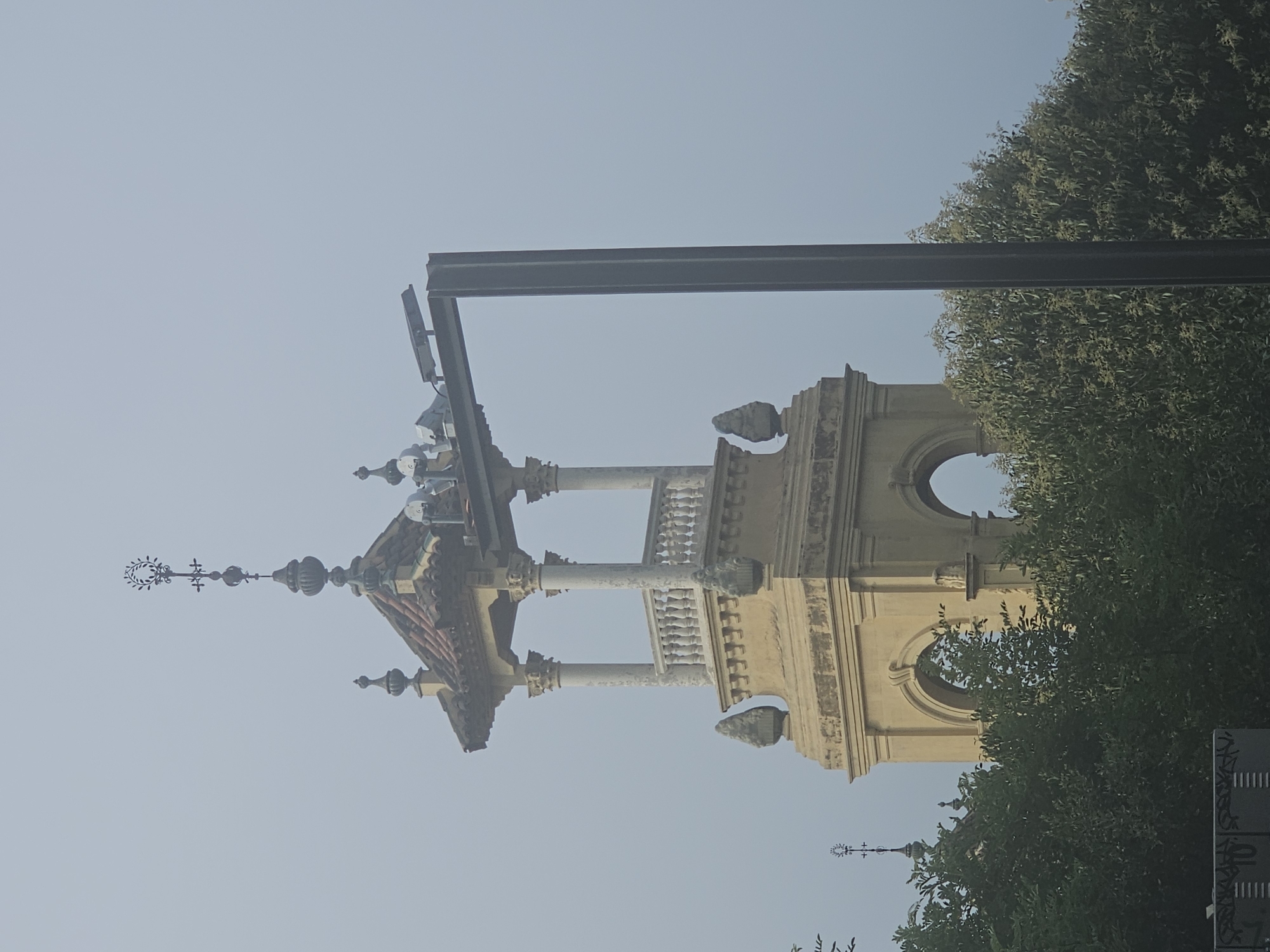 This image shows a view of a tower in Barcelona, Spain, partially obscured by trees and a lamppost. The tower has a classical architectural style, with arched openings and decorative elements. The sky is overcast, creating a soft, diffused light. A metal lamppost with a light fixture is positioned in the foreground, to the right of the tower. Several birds are perched on the structure supporting the lamppost. Trees and foliage are visible at the bottom of the image, with a portion of the tower visible above them. The overall impression is one of a historical building in an urban setting.
