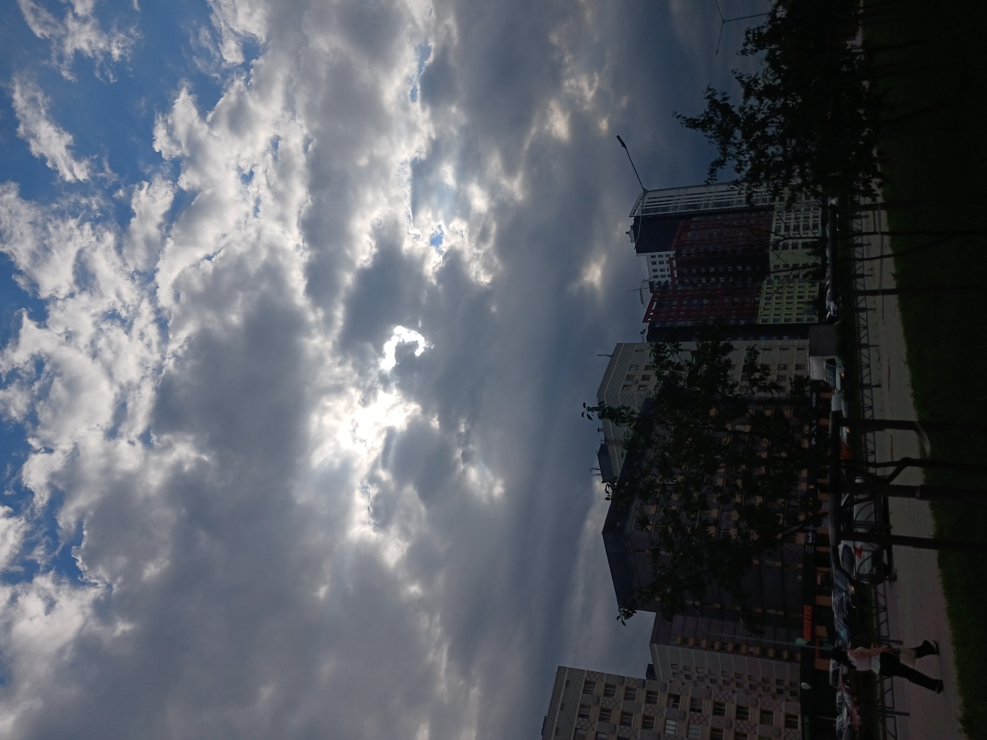 The image shows a residential area in Yekaterinburg, Russia, with several multi-story buildings visible against a dramatic sky. The buildings are modern and diverse in their architecture and color, with some featuring lighter facades and others darker, more industrial designs. Trees and streetlights line the foreground, casting shadows and adding to the urban landscape.
The sky is partly cloudy, with shafts of sunlight breaking through the dark, heavy clouds, creating a dynamic and somewhat ominous atmosphere. One of the buildings has signage that appears to be in Russian, including the word "MoreTka," which is a Russian word for a type of fish.
In the lower left of the image, a person is walking along a sidewalk, dressed in a white top and dark pants. They are carrying a bag. Cars are parked and driving on a street in the mid-ground, suggesting a typical urban scene. The overall impression is one of a cloudy day in a populated city, with a mix of modern architecture and natural elements.