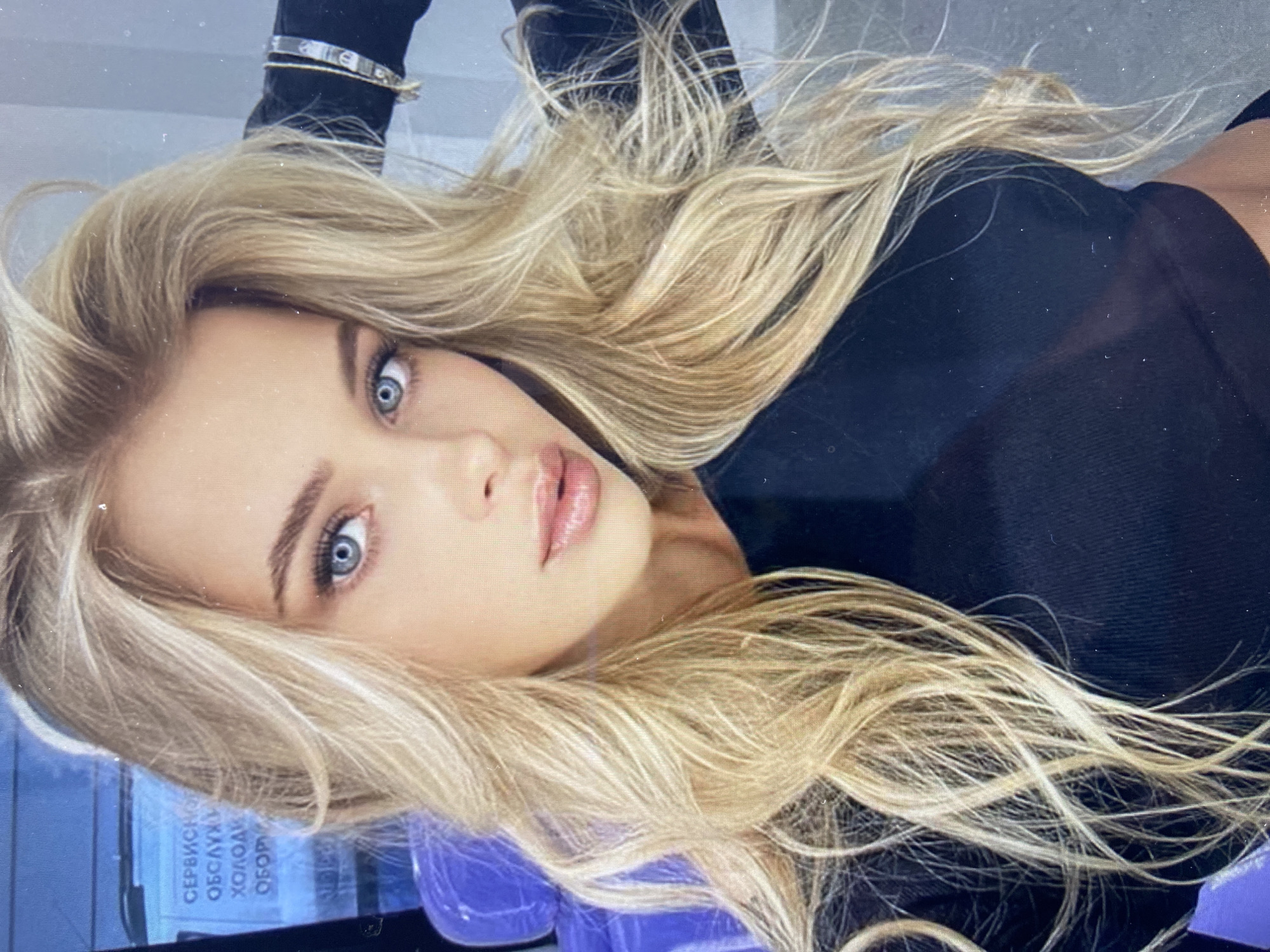 A close-up, head-and-shoulders studio portrait shows a young Caucasian woman with long, wavy blonde hair. She has striking blue eyes, fair skin, and full lips, which are slightly parted and covered in a glossy lipstick. Her makeup includes defined eyebrows and mascara. She is wearing a black, long-sleeved, fitted top. Her left arm is raised behind her head, with her hand resting on her hair. A glimpse of a purple chair is visible to the left, and a sign with Cyrillic lettering is partially visible in the background. The lighting is bright, casting soft shadows and highlighting her features. The overall impression is one of a posed, professional photograph.