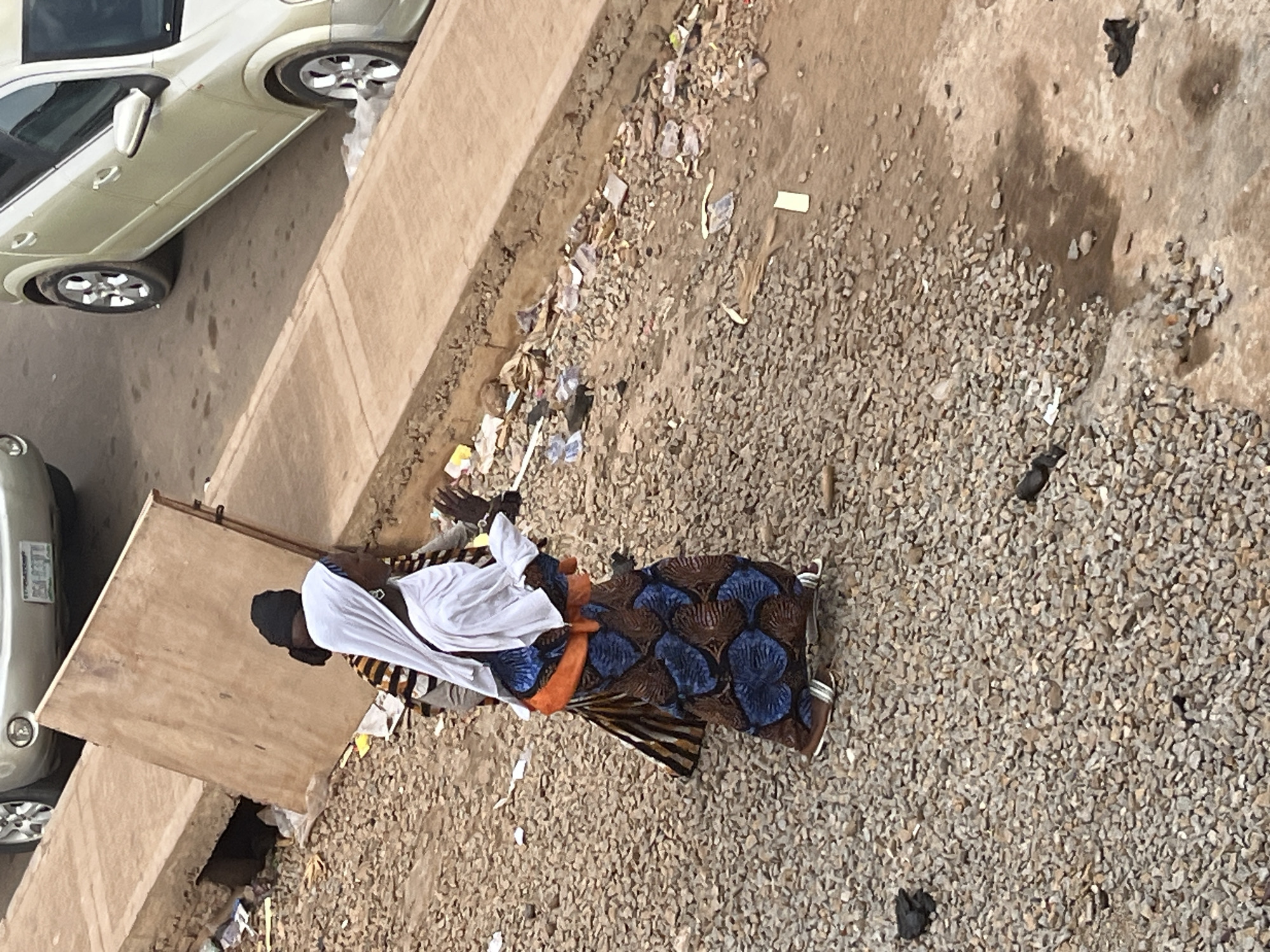 The image captures a street scene in Jos, Nigeria, with a focus on two individuals and their surroundings. In the foreground, a woman is walking, wearing a white hijab, an orange sash around her waist, and a blue and brown patterned dress. She has dark skin, wears silver sandals, and her left hand is raised, possibly holding something or gesturing. Behind her, another individual is partially visible, wearing a black hijab and a striped shirt.
The environment is characterized by a rough, gravelly surface. A concrete curb runs along the right side of the image, with a wooden board leaning against it in the background. The area appears to be a roadside, with some debris and scattered litter on the ground, including plastic wrappers and papers. Two light-colored cars are parked or driving in the background, one with its rear visible and the other partially visible from the side. The overall impression is of an urban outdoor setting in Jos, with a mix of pedestrian activity and vehicular presence.