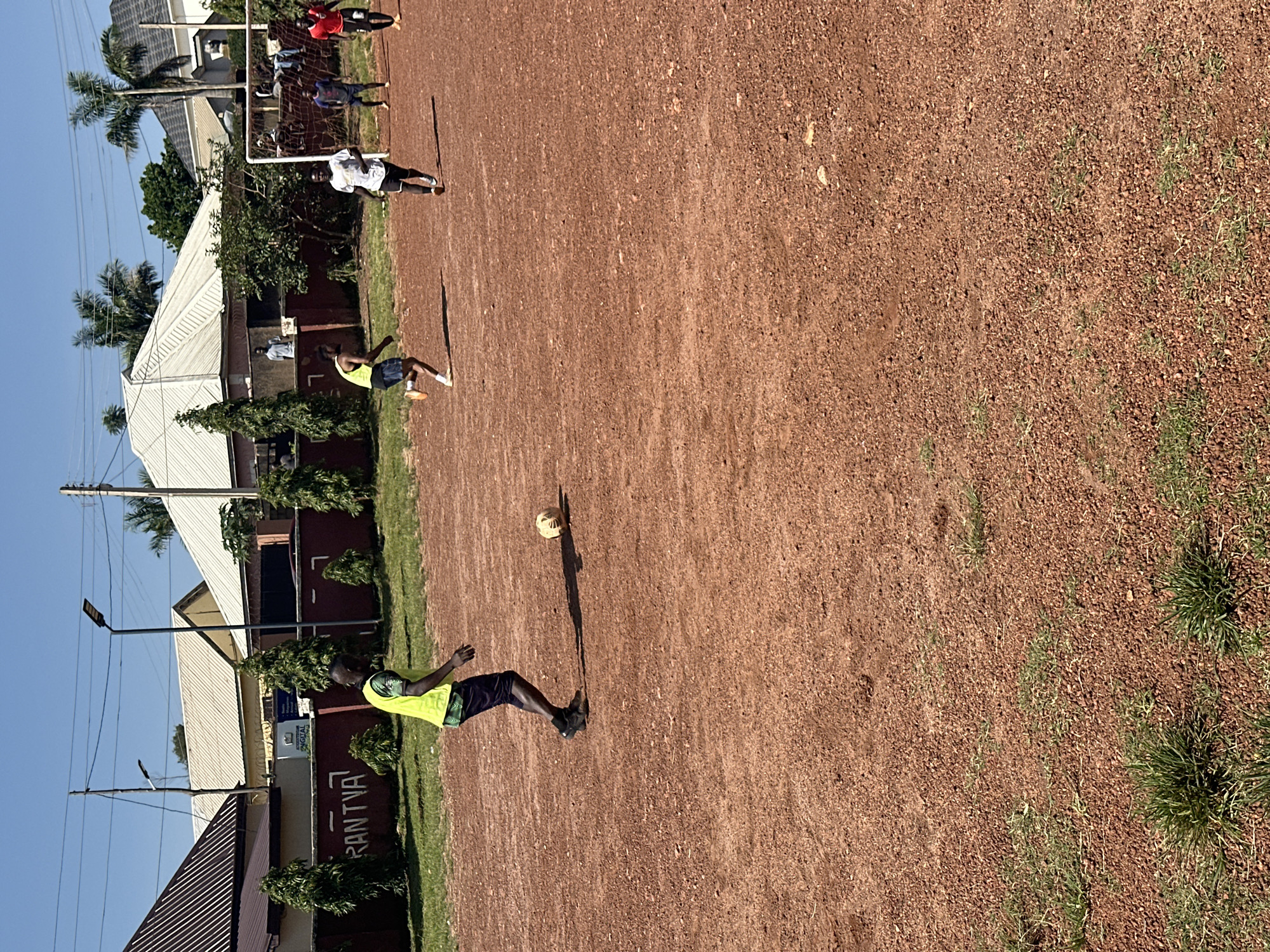 In an outdoor football field in Jos, Nigeria, five young men are engaged in a game. The field's surface is predominantly reddish-brown dirt, interspersed with patches of dry grass. The weather is clear and sunny. Two men are in the foreground, one wearing a bright yellow vest over dark shorts and black sneakers, actively playing football. The other, in a white t-shirt and dark shorts, is running towards the goal. In the background, three other men are visible; one in a blue t-shirt and shorts, another in a red t-shirt and black shorts, and a fifth individual further in the background. A football goal is set up on the right side of the frame, with a net. Behind the field, a brick wall with a sign that reads "RANTVA" is visible, along with several buildings, including houses with corrugated metal roofs and palm trees. Electrical poles and wires are also present in the background.