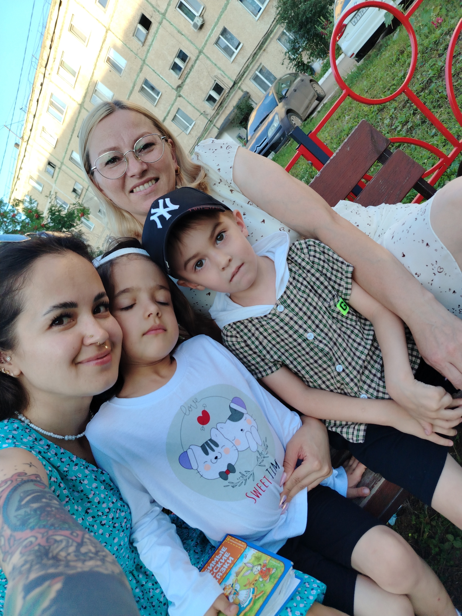A group of four people, two women and two children, are seated on a park bench outdoors. The woman in the foreground on the left, wearing a turquoise floral dress and a pearl necklace, has her left arm on her lap and is looking at the camera. Her arm has tattoos. Next to her, a young girl with dark hair and a white shirt with a cartoon cat print is leaning on her. To the right of the girl, a boy in a plaid shirt and a black baseball cap is looking to the side. Behind the children, a woman with blonde hair and glasses is smiling and looking at the camera. They are sitting on a wooden bench with a red metal frame. In the background, there is a light-colored apartment building, a dark blue car, and a white car. The ground is covered in grass and fallen leaves. The image was taken in Yanaul, Russia, likely in a public park or courtyard area.