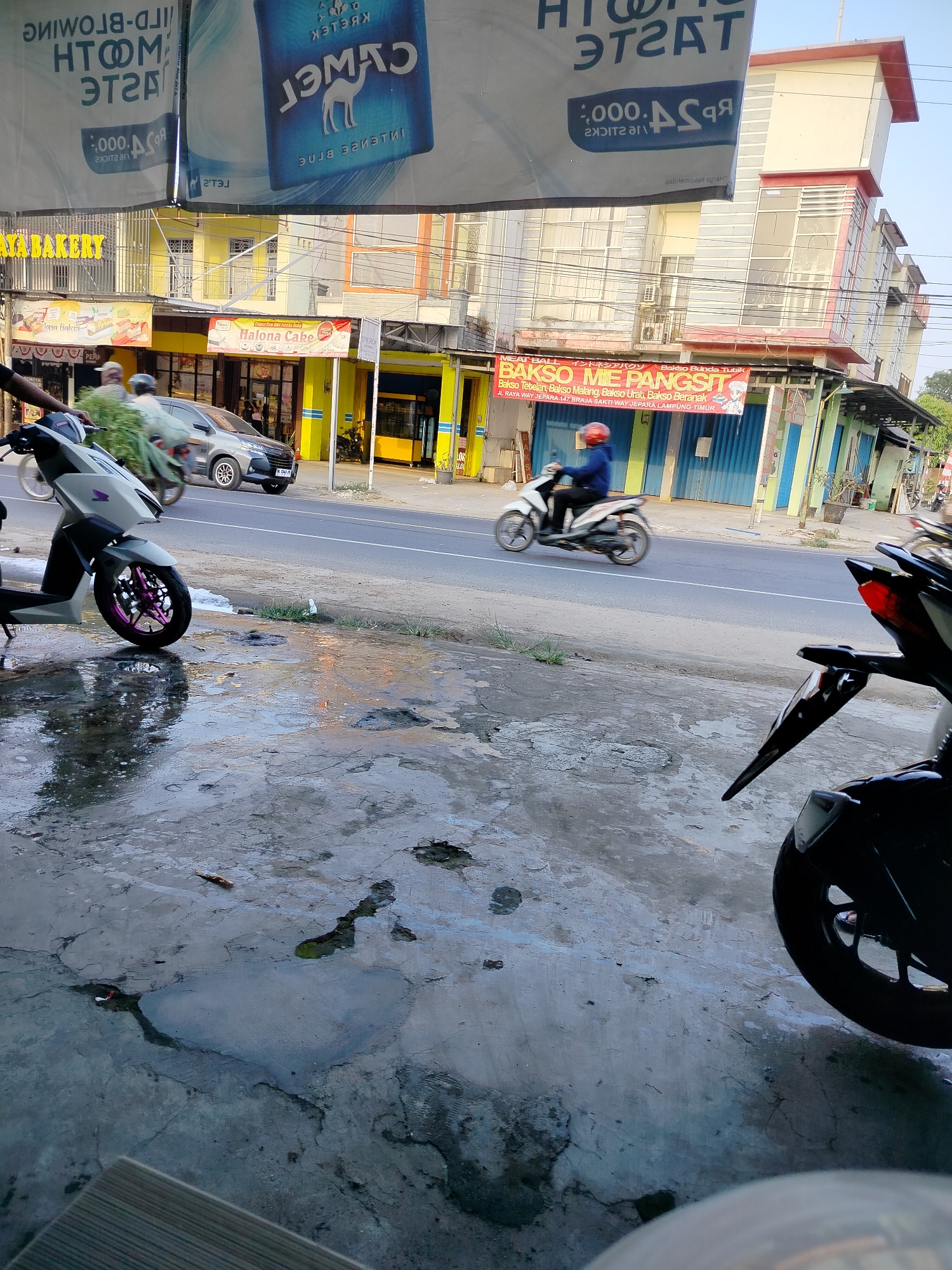 The image depicts a street scene in Braja Asri, Indonesia, on a sunny day. In the foreground, a light gray scooter with a pink rim on its front wheel is parked on a wet, cracked concrete surface. To the right, the rear of a black motorcycle is visible, with its taillight illuminated.

In the middle ground, a gray car is driving on a paved road. Beside the road, a man on a white scooter, wearing a red helmet and a blue jacket, is riding past a row of buildings. The buildings feature various storefronts, including one with a yellow sign reading "JAYA BAKERY" and another with a red banner advertising "BAKSO ME PANGSIT". The buildings have multi-story facades with windows and some appear to be undergoing construction or renovation.

The background shows more buildings and trees under a clear blue sky. The overall impression is a bustling urban environment with ongoing commercial activity.