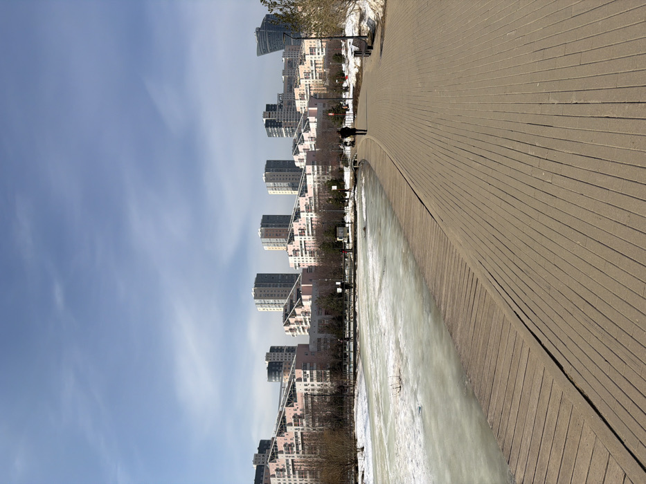 This image depicts an urban park scene in the Setun River Valley, Kuntsevo district, Moscow, Russia, during what appears to be late winter or early spring.
In the foreground, a wide, light-colored paved pathway (likely concrete pavers) extends from the lower right, curving gently to the right as it recedes into the midground. Parallel to this pathway, on the left, a narrower wooden boardwalk runs alongside a body of water. The water is approximately one-third covered in a layer of ice and slush.
In the midground, one individual, dressed in a dark coat, walks away from the viewer on the paved pathway. Further ahead on the same path, a second, lighter-clad individual is visible, possibly pushing a stroller or pram. Bare deciduous trees and some evergreen shrubs line the edges of the pathway and the opposite bank of the water. Patches of residual snow are visible along the path's edge and around the trees. Several park benches and streetlights are spaced along the paved promenade. Small bridges span the waterway in the distance.
The background is dominated by a distinctive urban skyline. A series of residential complexes feature multi-story buildings with prominent pink, sloped terraced roofs and light-colored facades. Behind these, several taller, grey and white rectangular apartment blocks are visible. On the far right, a modern, tall building with a unique curved glass facade stands out. The sky above is clear blue with minimal, wispy clouds.