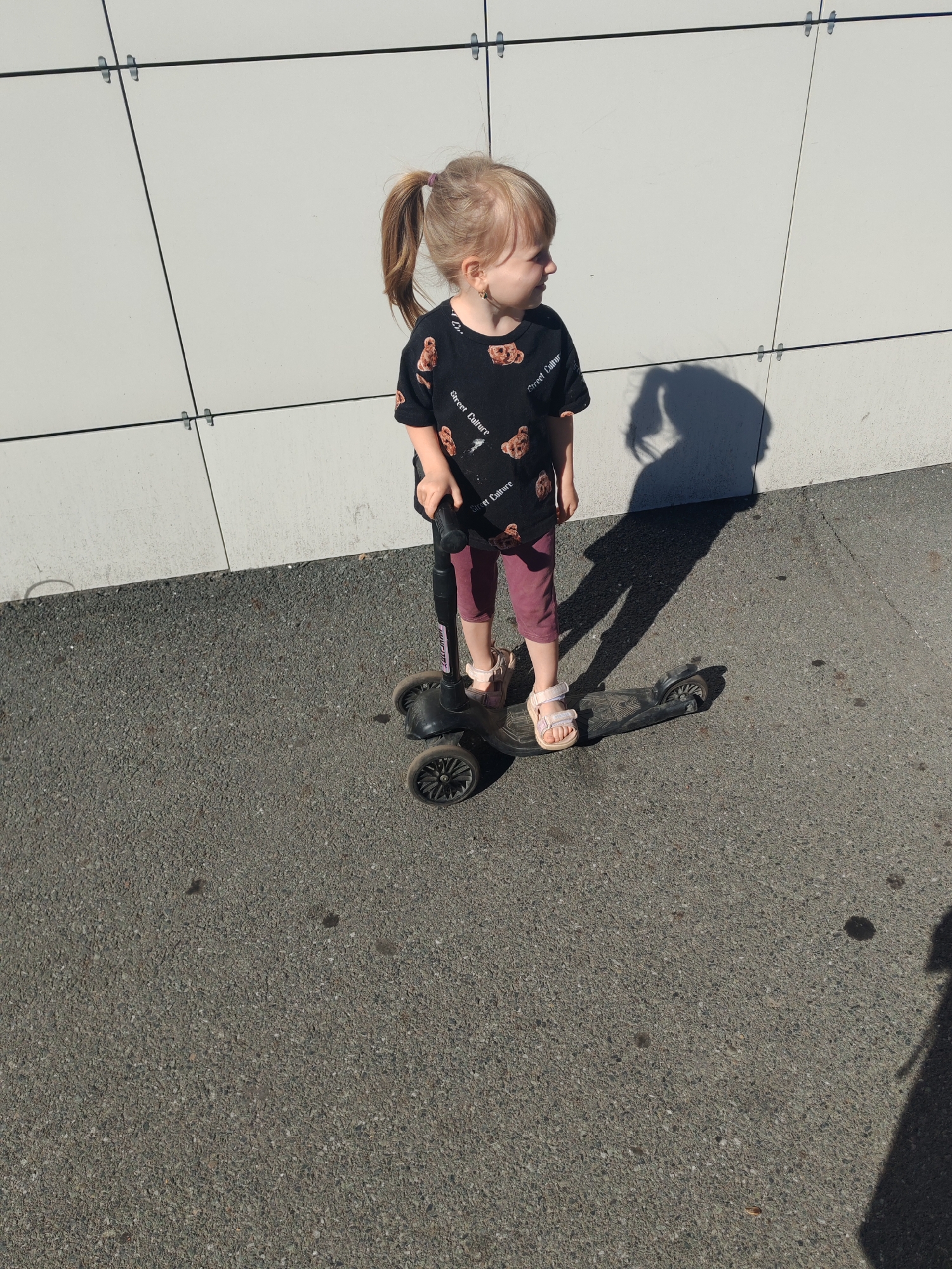 A young girl with blonde hair in a ponytail stands on a scooter on an asphalt surface. She is wearing a black t-shirt with a teddy bear print and burgundy leggings. She is holding the handlebars of the scooter with her right hand. Her shadow is cast on the light-colored wall behind her. The wall is made of large square panels. The image appears to have been taken outdoors on a sunny day. The setting is a city street.