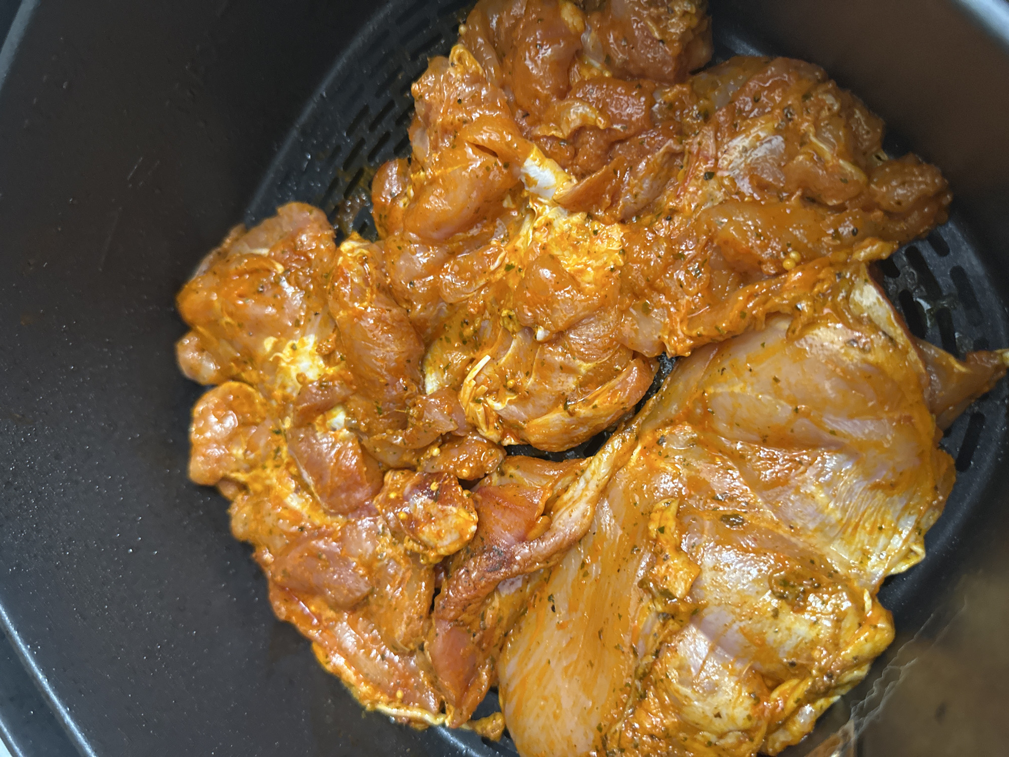 The media file presents a close-up, top-down view of raw, marinated chicken pieces within a black air fryer basket.
The primary subject is a generous quantity of chicken, cut into various, irregularly shaped pieces, consistent with boneless chicken thighs or similar cuts. Each piece is thoroughly coated in a vibrant, orange-red marinade. This marinade appears to be rich in spices, with visible specks of green herbs and darker granules, likely black pepper, indicating a flavorful preparation. The raw chicken beneath the marinade shows a pale pinkish-white color in areas where the coating is thinner.
The chicken is contained within a black, perforated basket, clearly identifiable as part of an air fryer. The grid-like structure of the basket's bottom and sides is visible, designed to facilitate hot air circulation during cooking. The material appears to be non-stick.
The image captures the stage of food preparation where marinated chicken is ready to be cooked. No people are visible, and therefore no emotions can be inferred. There is no visible text in the image. The time of day and weather conditions cannot be determined from the indoor setting. While the location context is provided as Sofia, Bulgaria, there are no visual cues within the image itself to corroborate this geographical detail. The scene is focused entirely on the culinary ingredients and equipment.