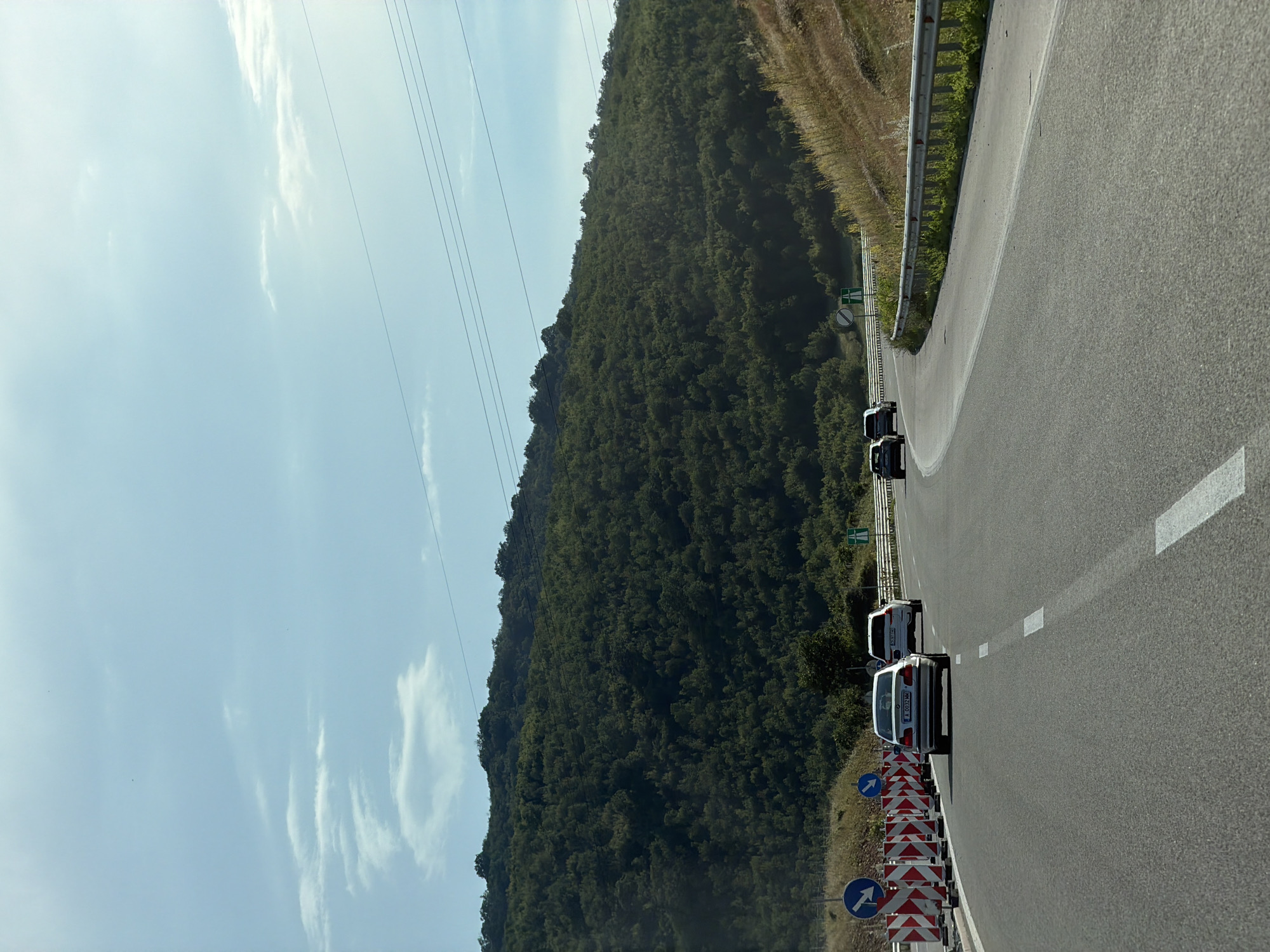 The image shows a view from a car driving on a highway near Brestnitsa, Bulgaria. The highway is asphalt with white lane markings. There are several cars on the road, including a silver BMW in the foreground and a white SUV behind it. To the left of the road, there are construction barriers and traffic signs. In the background, there is a densely forested hill under a bright blue sky with scattered clouds. Power lines are visible stretching across the sky. The road appears to be curving to the right. The scene depicts a typical daytime highway drive through a rural or semi-rural landscape.