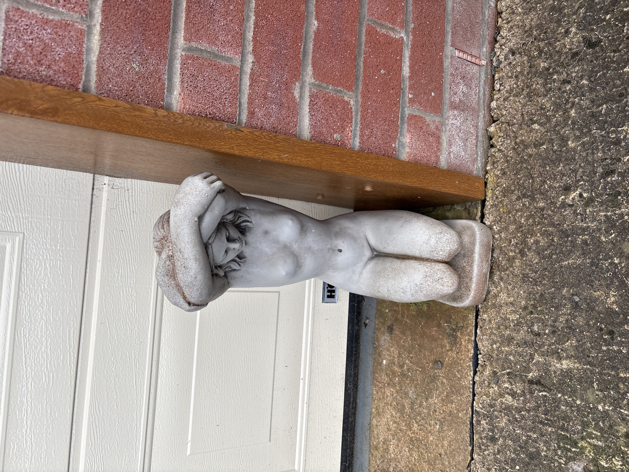 A supine stone sculpture of a human figure is positioned near a white door with a brown wooden lintel. A brick wall is above the lintel. The statue rests on a gray concrete surface. A black nameplate is affixed to the door below the statue. The location is Shrewsbury, United Kingdom.