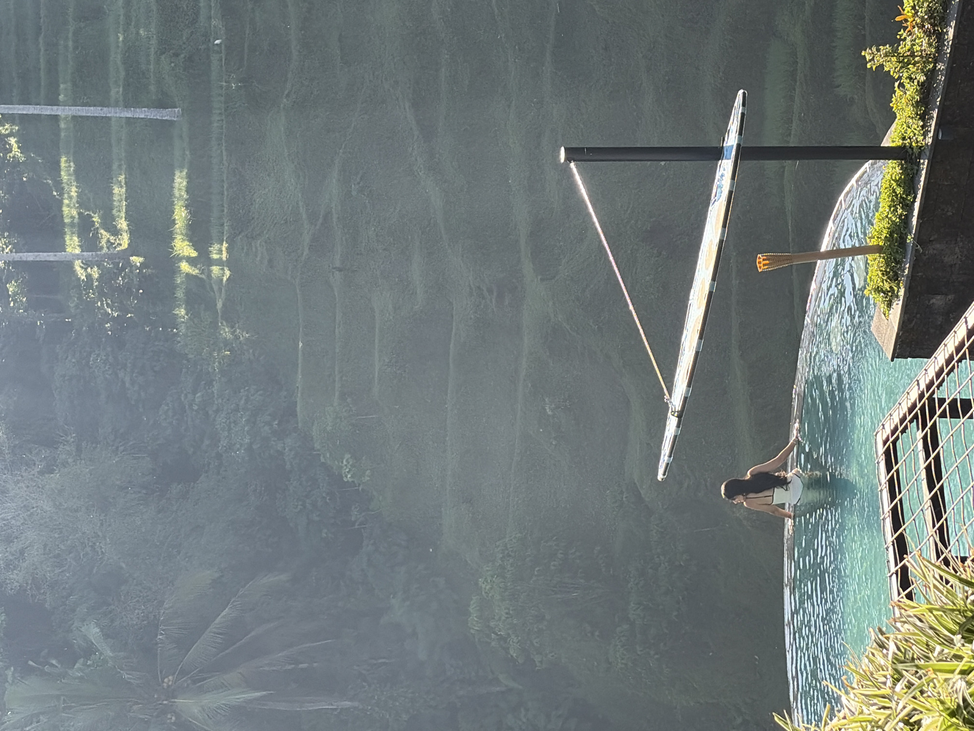 A woman is partially submerged in an infinity pool, her back to the camera. She is wearing a white swimsuit with black trim. The pool overlooks terraced rice paddies, a common sight in Tegallalang, Indonesia. The sun casts dramatic shadows across the landscape, highlighting the contours of the paddies. A tiki torch and a partially visible umbrella or canopy are positioned near the edge of the pool. The environment is lush and green, with trees visible in the background.