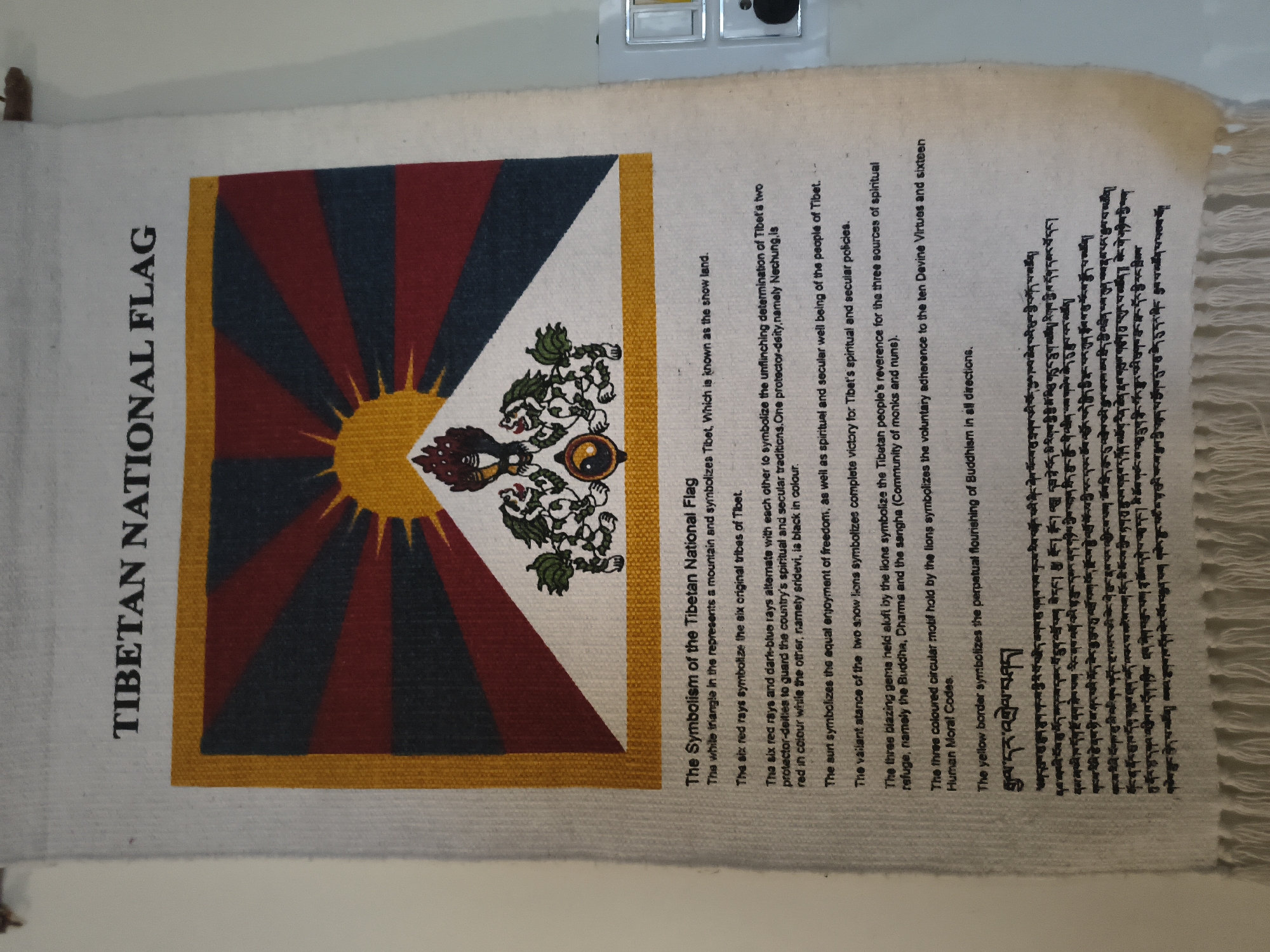 The image shows a Tibetan National Flag tapestry hanging on a wall. The tapestry displays the flag's design, which includes a sun, mountains, lions, and a Dharma wheel. Below the flag, there is text explaining its symbolism in English, followed by Tibetan script. The wall appears to be painted a light color, and a white electrical outlet with a switch is visible on the right side of the image. The overall setting seems to be indoors, possibly in a dwelling or a cultural center, with the tapestry serving as a decorative or informational item. There is no indication of the city of Haini, India, in the image or its context.