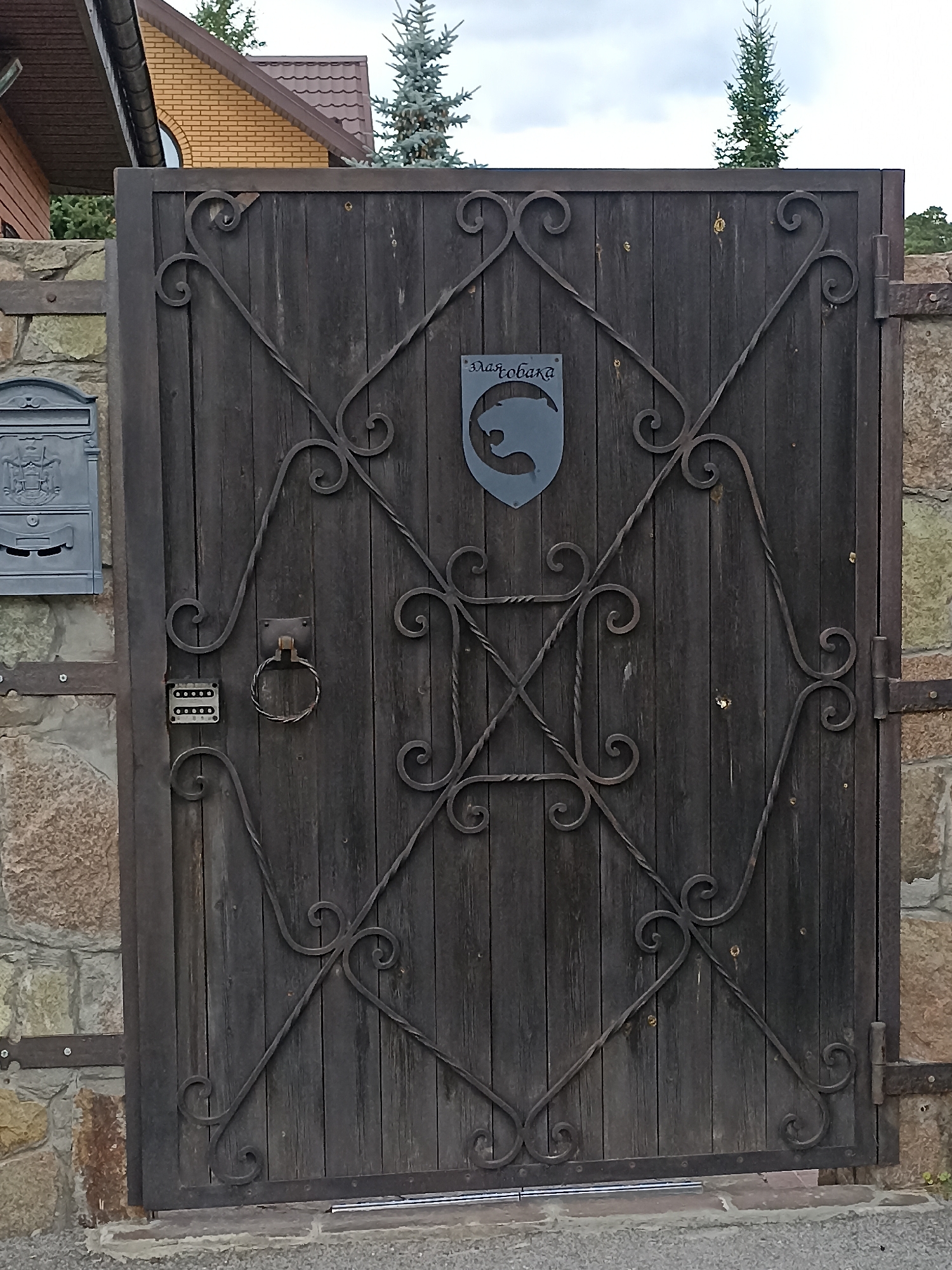 The image shows a dark wooden gate with ornate metalwork. On the gate, there is a shield-shaped emblem with a silhouette of a dog's head and the word "Злаясобака" (meaning "Angry Dog" in Russian) written above it. To the left of the gate, there is a stone wall and a grey metal mailbox. Below the mailbox, there is a keypad with buttons for entering a code. A circular metal ring handle is attached to the gate. The gate is part of a property, with a portion of a house and trees visible in the background. This property is located in the city of Mochische, Russia.