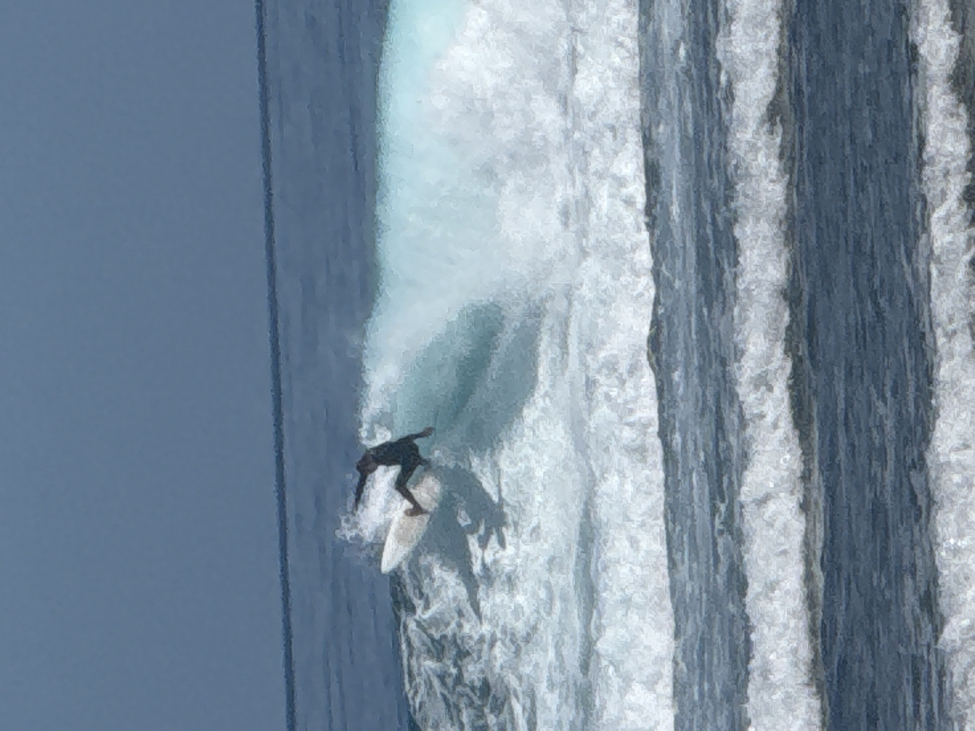 A surfer wearing a dark wetsuit is riding a white surfboard down the face of a large wave. The wave is breaking, with white foam and spray visible. The sky is a pale blue. The location is likely the Uluwatu surf break in Kuta Selatan, Indonesia.