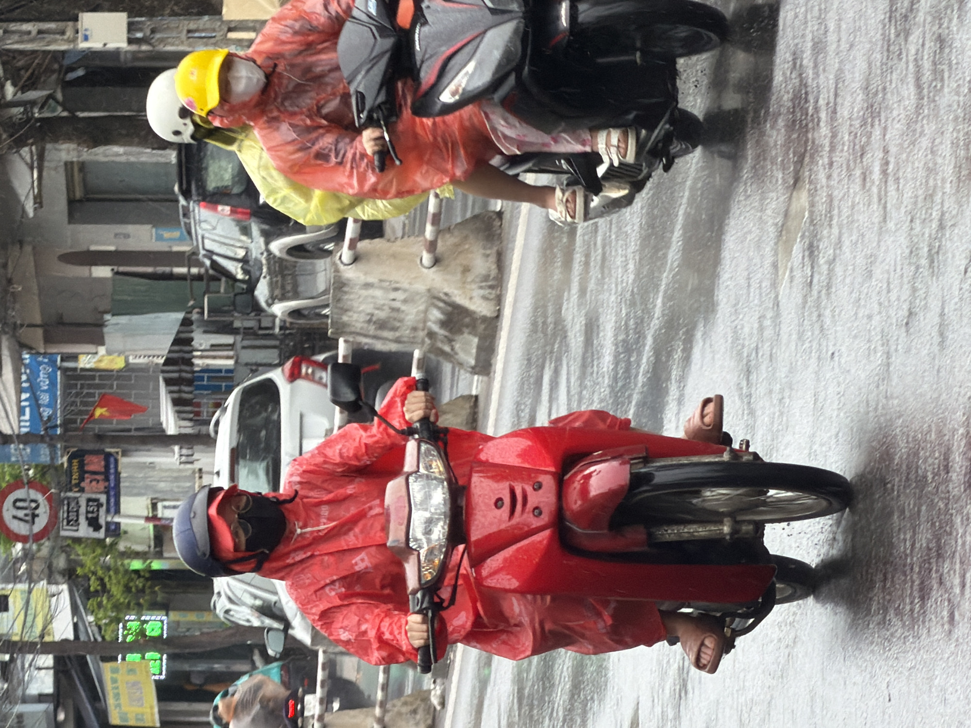 Two individuals on motorbikes are navigating a wet street in Nha Trang, Vietnam, under rainy conditions. The person in the foreground rides a red motorbike and wears a bright red rain suit, a dark helmet, and glasses. The individual behind them is on a black motorbike, also in rain gear, with a yellow helmet and white mask. A dark-colored SUV is visible in the background, along with various buildings and street signs, including a circular sign with "40" and a rectangular sign with Vietnamese text. The wet asphalt reflects the surrounding lights and scene.
