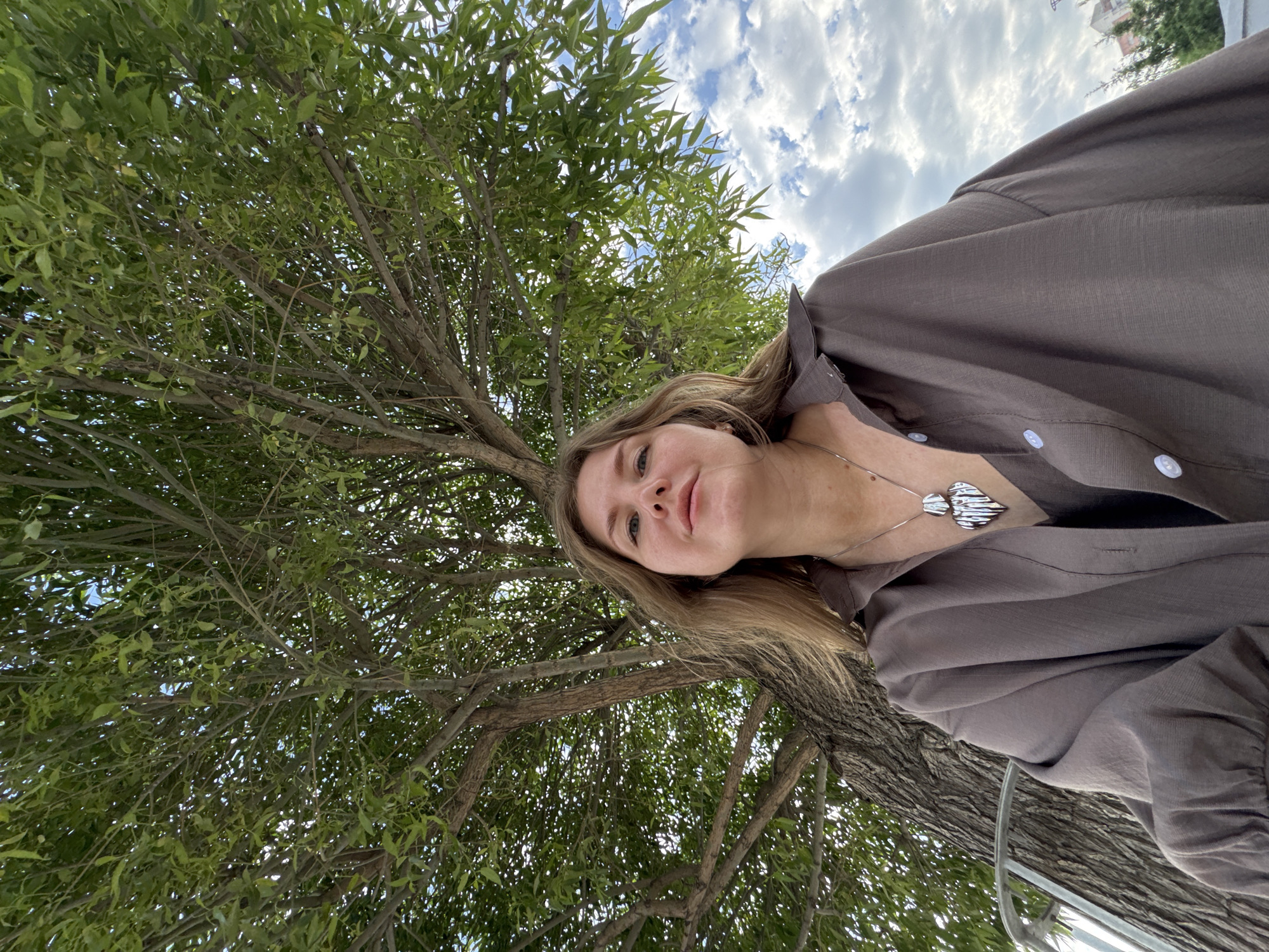 A low-angle, slightly askew photograph shows a light-skinned woman standing underneath a large tree. The tree fills a significant portion of the left side of the frame, with its branches extending upwards. Some patches of sky and clouds are visible through the foliage. The woman is positioned on the right side of the image, facing the camera with her head tilted slightly. She has blonde hair, and is wearing a brown button-down shirt and a silver necklace. The location is Perm, Russia.
