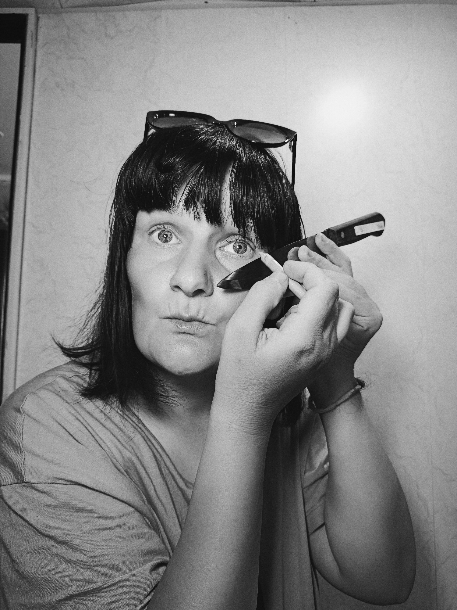 A black and white medium shot depicts a woman holding two objects in her hands, positioned near her face. The woman's facial expression includes slightly pursed lips and wide eyes, suggesting surprise or concentration. Sunglasses are perched atop her head, partially obscuring her hair. Her dark hair is cut in a style with bangs. The objects she holds include what appears to be a knife and a lighter. The background is a plain wall. The city and specific location are not evident from the image.