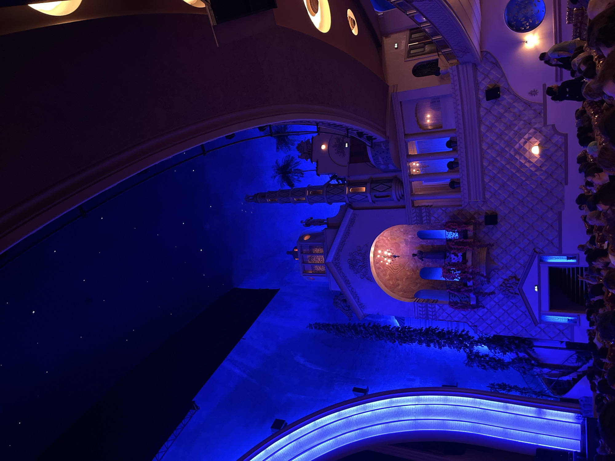 The image shows the interior of the Paradis Latin cabaret in Paris, France. The scene is illuminated with predominantly blue and purple lighting, with some warm orange light sources also visible.
In the foreground, a crowd of people are seated, appearing to be an audience, facing away from the camera. The seating arrangement is typical of a theater or cabaret.
The mid-ground and background depict the ornate architecture of the Paradis Latin. The ceiling is a deep blue with small white lights that resemble stars, creating a night sky effect. To the left, a large, curved structure with vertical blue LED lighting dominates the space. To the right of this, a multi-tiered building facade is visible, designed to evoke an exotic or fantastical setting. This facade features architectural elements such as arches, balconies, columns, and decorative textured walls. There are also palm tree motifs and what appear to be statues or figures integrated into the architecture.
The overall impression is a vibrant and theatrical atmosphere, characteristic of a performance venue.