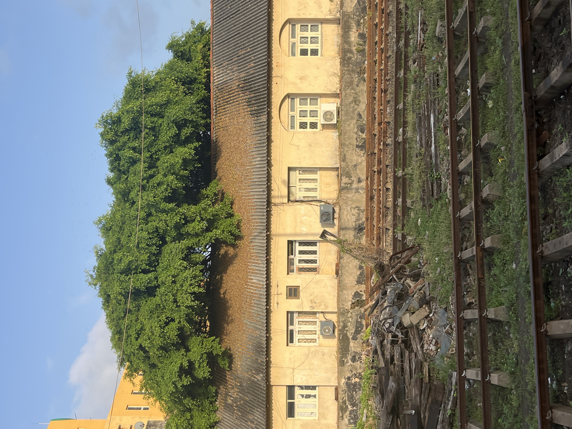 The image depicts a weathered, single-story building with a corrugated roof, partially obscured by a large, lush green tree. The building's facade is painted a pale yellow, with several windows featuring latticed panes. Air conditioning units are visible beneath some of the windows. In front of the building, there is a jumble of debris, including wooden planks, bricks, and metal scraps, situated near a series of train tracks. The train tracks extend into the foreground, with some appearing overgrown with grass. In the background, a bright yellow multi-story building is partially visible. The sky is a clear blue with a few wisps of clouds. This scene appears to be located in Galle, Sri Lanka, likely in an area near the railway station, characterized by its colonial-era architecture and the presence of both active and disused railway infrastructure.