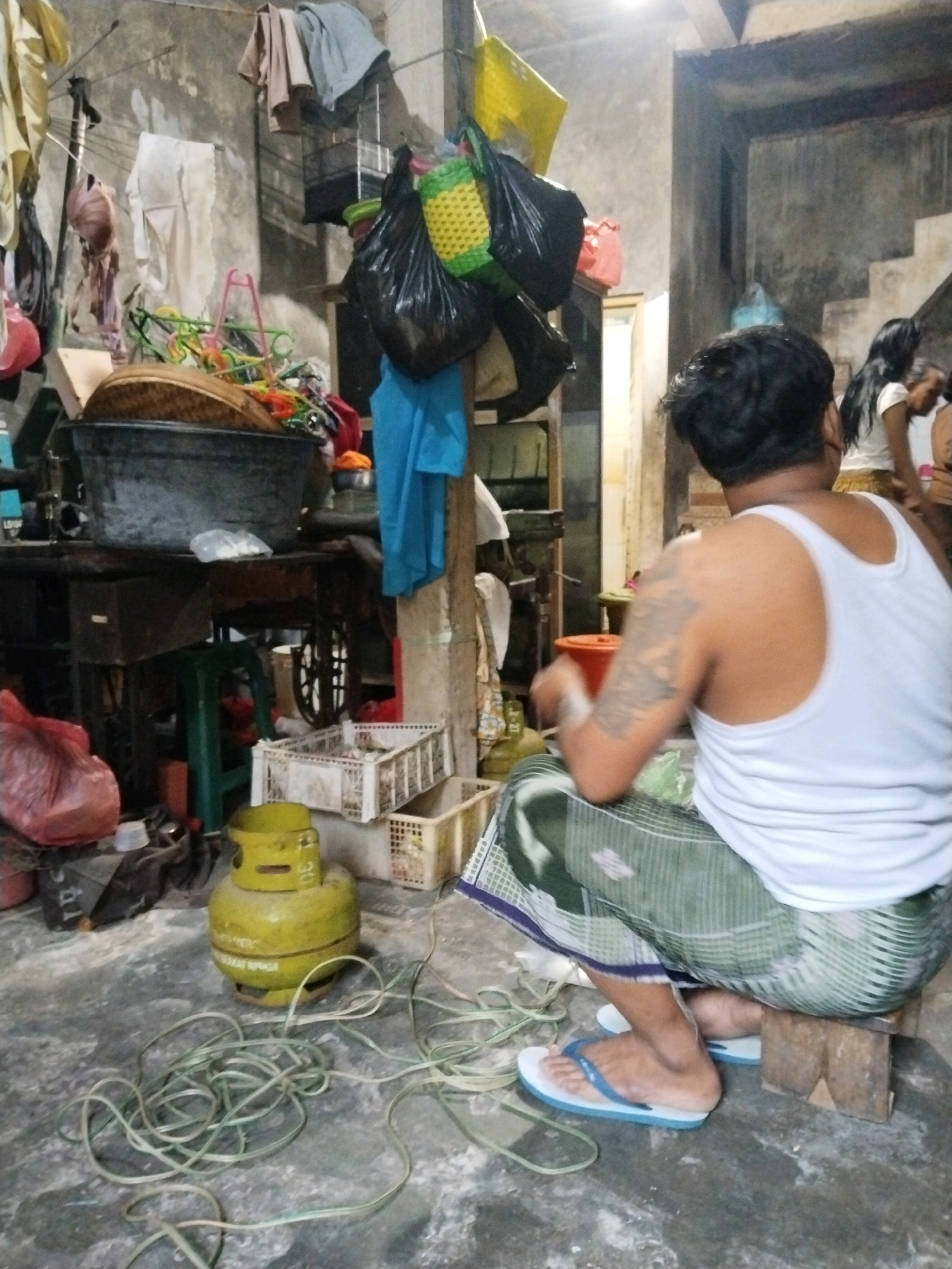 The image depicts an interior scene of what appears to be a cluttered and somewhat derelict dwelling in Pepelegi, Indonesia. In the foreground, a man wearing a white tank top and a green and white patterned sarong is seated on a small wooden block, facing away from the camera. His left arm is tattooed. He is wearing blue flip-flops. In front of him, a yellow gas cylinder sits on the concrete floor, with a green cable coiled around it.
The environment is characterized by a rough, concrete floor and walls. Various objects are scattered around, including plastic bags, baskets, and what looks like laundry hanging on lines. A large black basin is prominent on a wooden table behind the man, filled with a woven item. In the background, a staircase is visible, and a woman with long dark hair can be seen standing near it. The overall impression is one of a very lived-in and unkempt space.