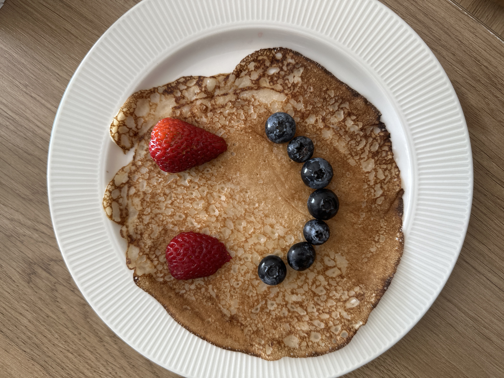 A close-up, top-down shot shows a single, round, golden-brown pancake arranged on a white, ribbed plate. The pancake has a slightly irregular edge, suggesting it was cooked in a pan. Two bright red strawberries are placed at the top of the pancake, acting as eyes, and a curved line of dark blue blueberries forms a smile below them. The plate rests on a light brown wooden surface. The scene is illuminated from above, casting soft shadows. The image is an overhead view of food art. The location is not determinable from the visual information provided in the image.