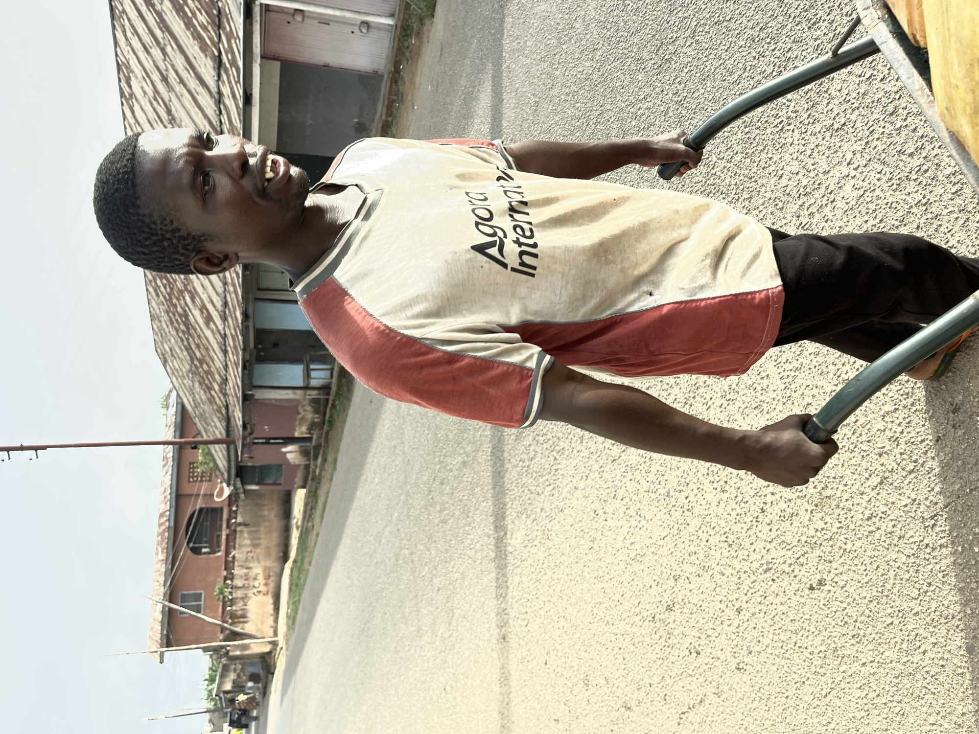 The image shows a man pulling a wheelbarrow on a paved road. He is wearing a light-colored t-shirt with "Agora International" printed on it, along with red accents on the sleeves, and black pants. The road surface is light beige with a rough texture. In the background, there is a building with a weathered roof and what appears to be another building further down the road. The sky is overcast. The location appears to be in a neighborhood in Jos, Nigeria.