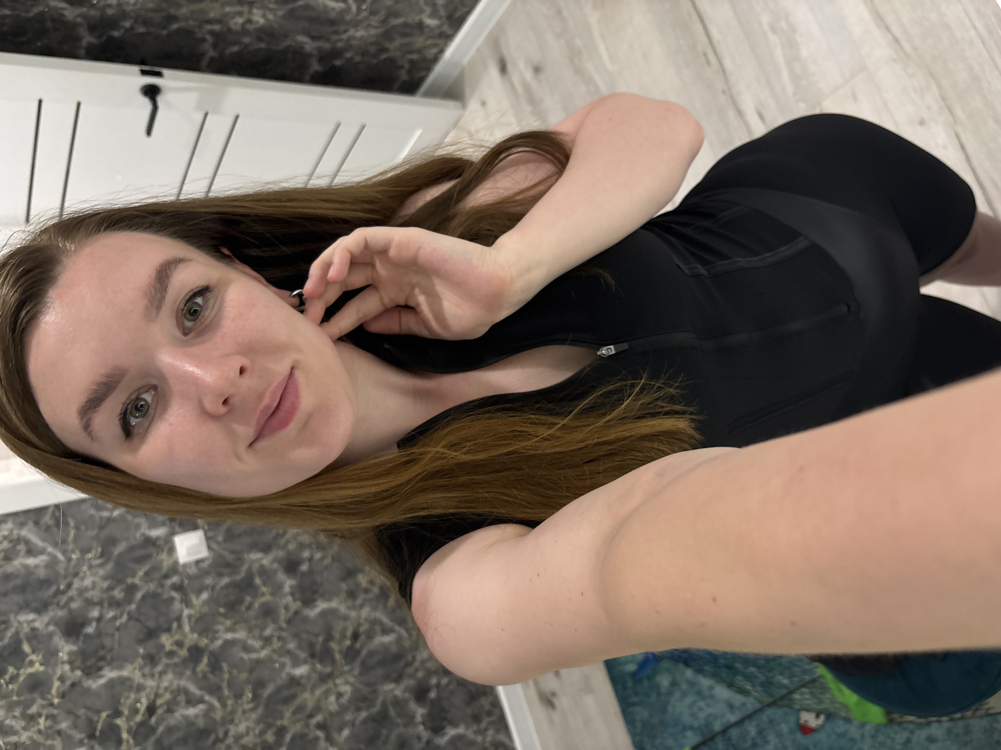 A selfie taken by a woman with long brown hair, wearing a black athletic outfit, possibly in an indoor location. The background includes a gray patterned wall and a white door. There is no information in the image to suggest this image was taken in Izhevsk, Russia.