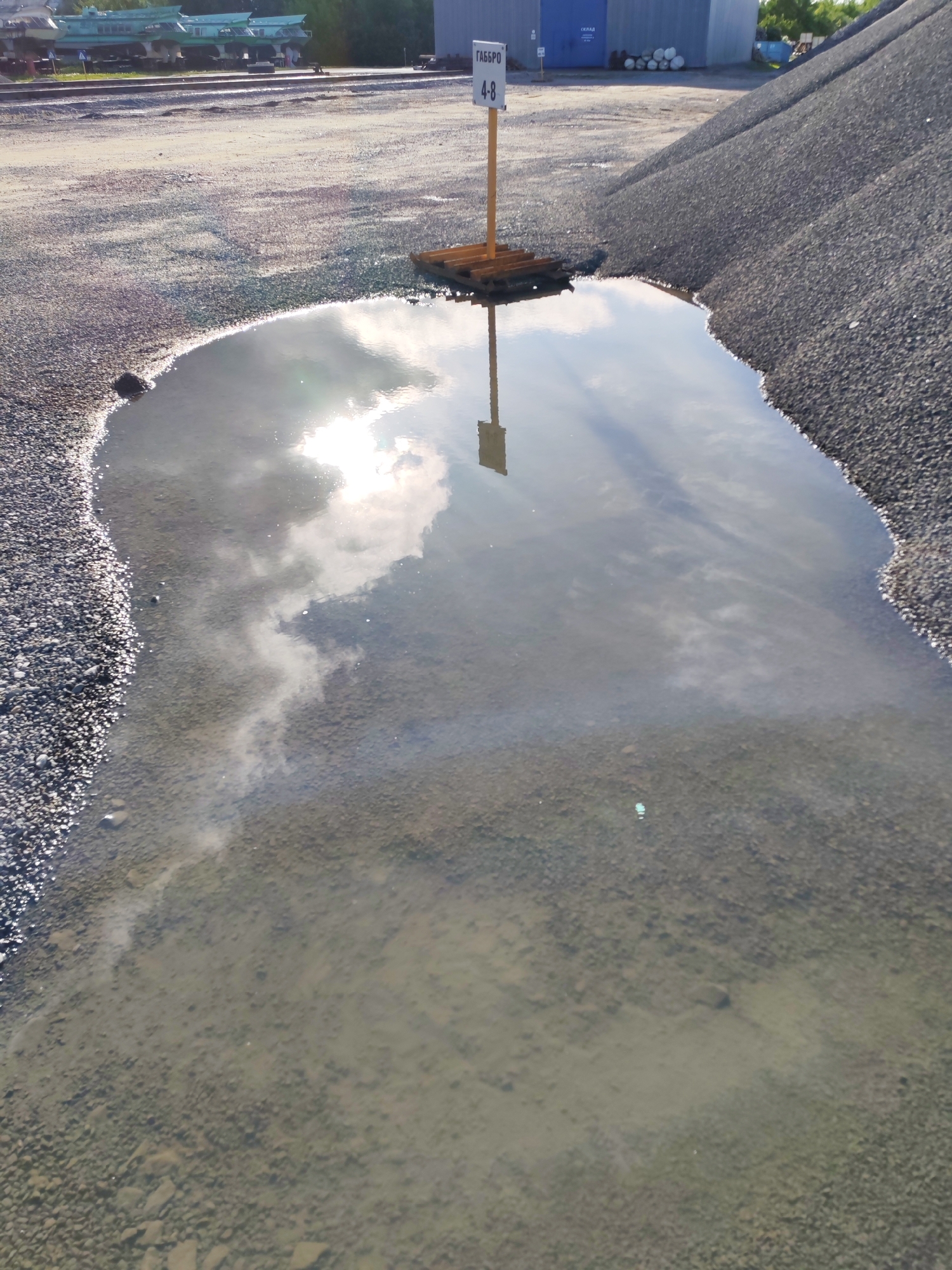 The image depicts a puddle of water on a gravel surface in an outdoor industrial area, with a sign reading "ГАББРО 4-8" (Gabbro 4-8) standing in the center. The puddle reflects the sky, including clouds and sunlight. To the left, a train car sits on a train track. Behind the sign, there is a large pile of gravel on the right, and in the background, there is a blue building, several rolls of white material, and some greenery.