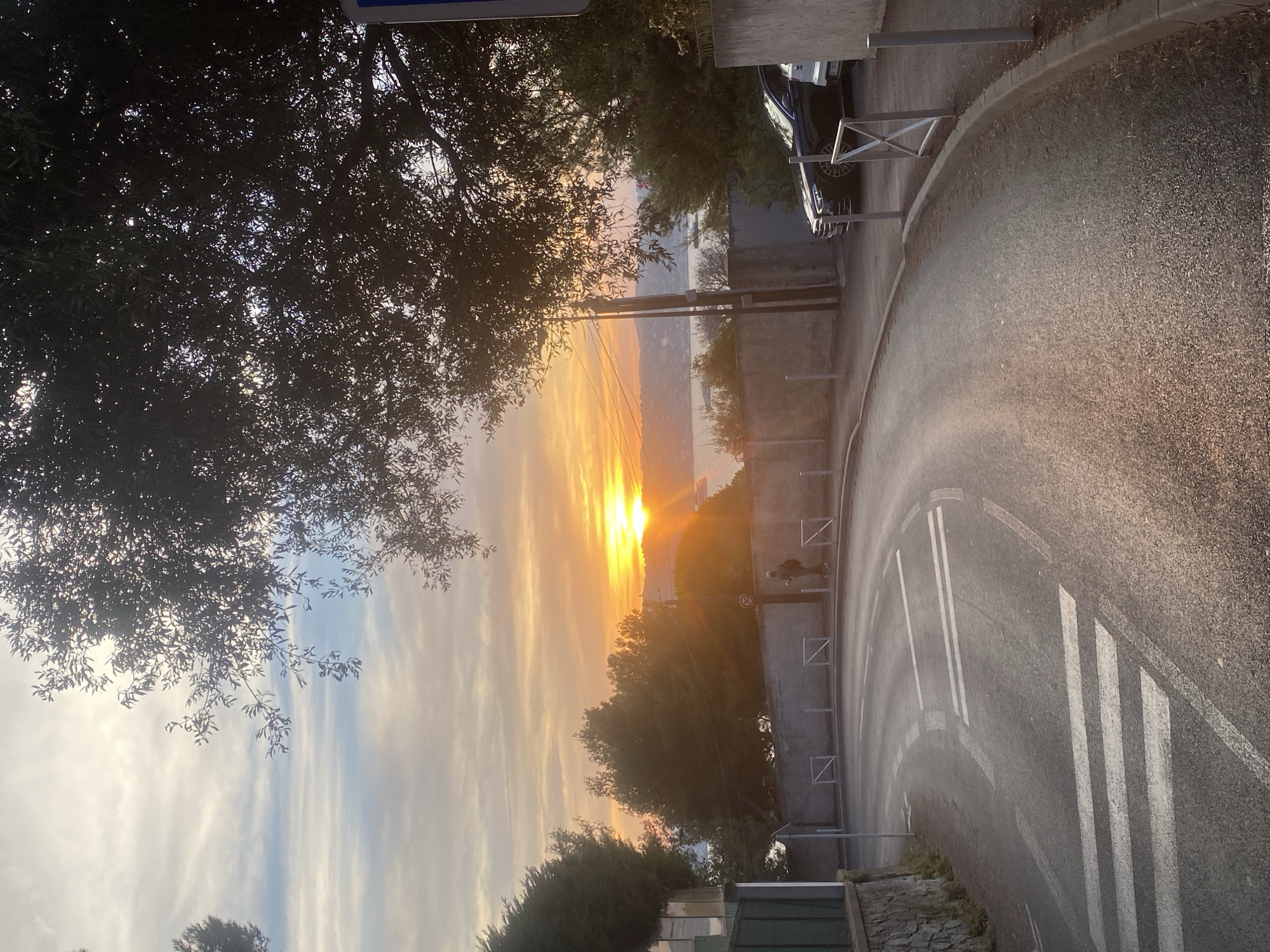 The image depicts a street scene in Antibes, France, during a sunset. A winding road with visible pedestrian crossings is in the foreground, leading towards a bay with boats in the distance. The sky is filled with warm hues of orange and yellow, characteristic of a sunset, with the sun casting a bright glow over the water. Trees, silhouetted against the sky, frame the upper portion of the image. A person is walking on the sidewalk, and several cars are parked along the street. The environment appears to be a coastal town with residential buildings and walls bordering the road.