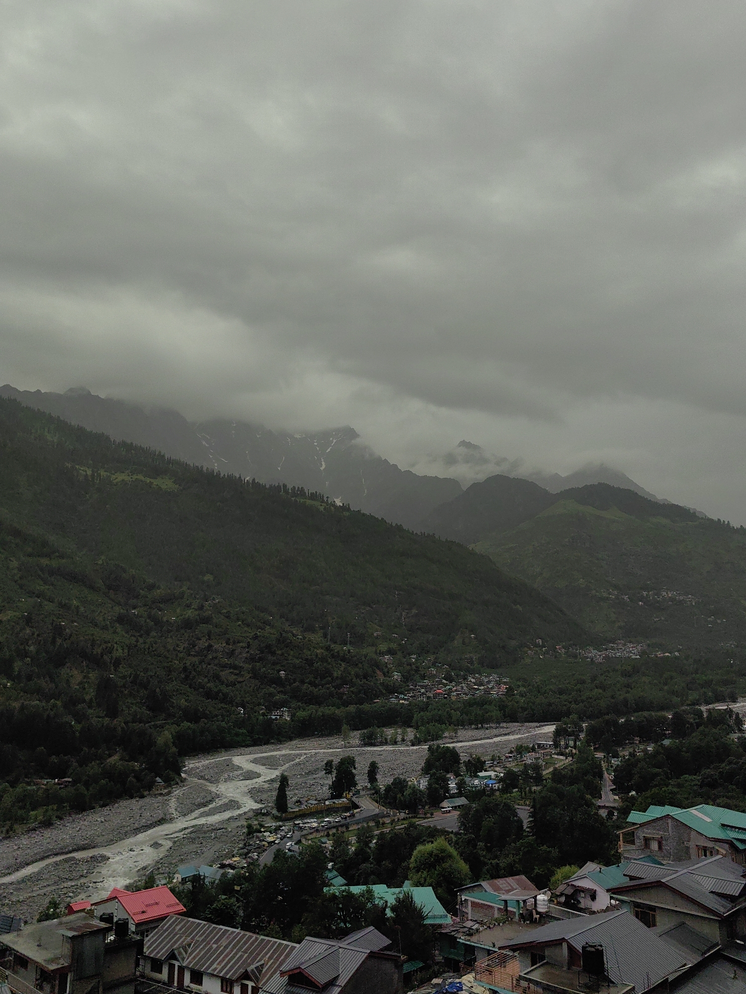 This is an aerial, overcast view of a mountainous, forested area with a village and river in Vashisht, India. The sky is a uniform grey, filled with thick clouds that obscure the tops of the mountains in the distance. These mountains are covered in dense, dark green vegetation, with patches of snow or lighter-colored rock visible on some of the higher peaks.
In the mid-ground, a wide, rocky riverbed meanders through the valley. The water appears somewhat murky, with a light brown or grey hue. Along the banks of the river and extending into the valley are clusters of buildings, forming the village. The architecture is varied, with some houses featuring bright red or green roofs, while others have darker, metal roofs. Some buildings are made of stone or concrete. There are numerous trees interspersed throughout the village and surrounding areas. A road with some parked vehicles is visible near the river.
In the foreground, more rooftops are visible, indicating a closer perspective of the village settlement. The overall impression is one of a somewhat subdued, natural landscape under a heavy, cloudy sky, with human settlements integrated into the valley. No people are clearly discernible in the image.