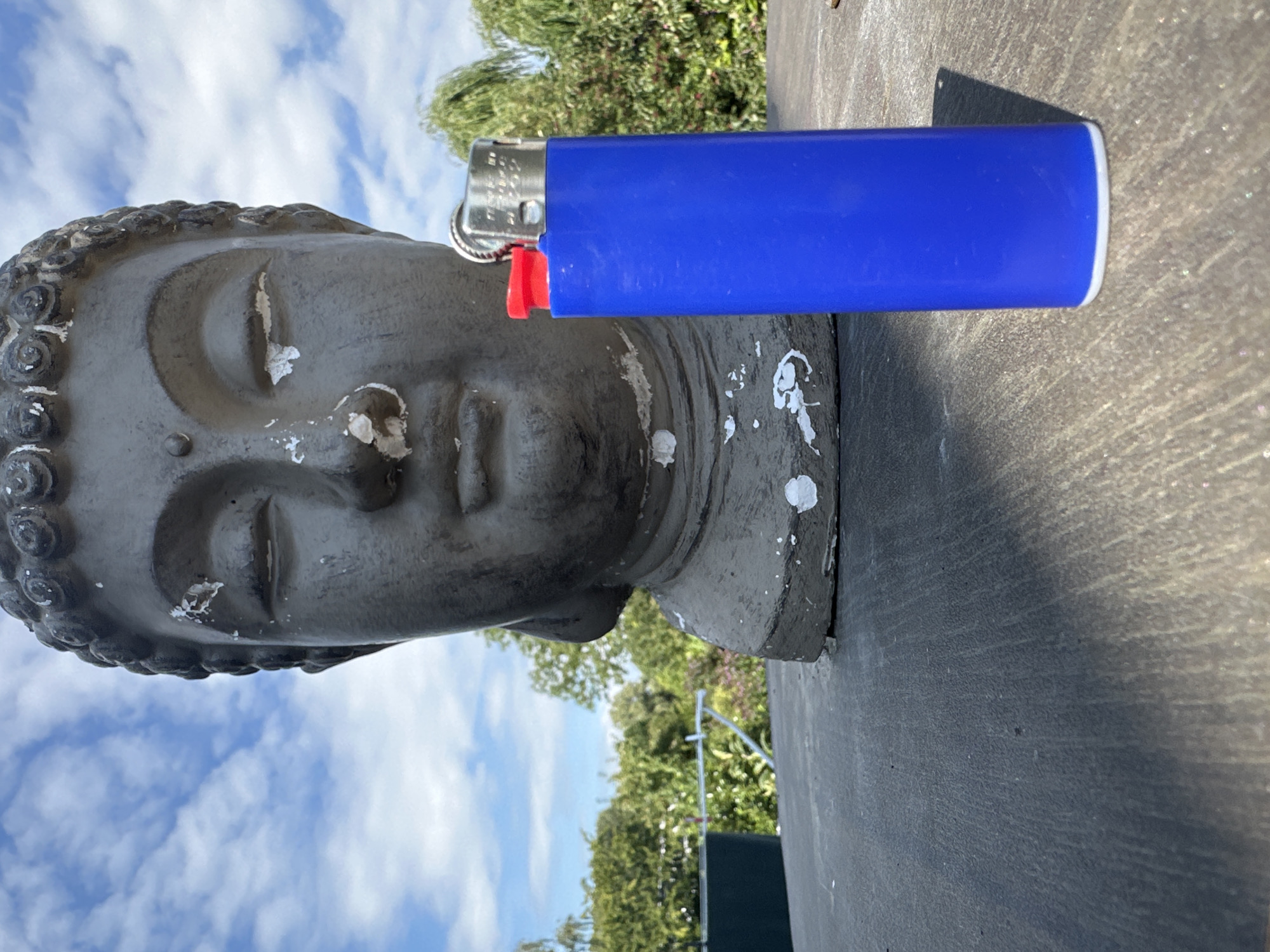 A close-up, low-angle shot captures a weathered, dark gray Buddha head statue situated behind a bright blue disposable lighter with a red plastic trigger. The Buddha's eyes are closed, and its face shows signs of wear and tear, with chips and white paint flecks on its surface, particularly around the eyes and forehead. The lighter is positioned prominently in the foreground to the right of the statue.

The background reveals a lush, green outdoor environment under a bright blue sky dotted with white, fluffy clouds. Dense foliage from trees and bushes forms a natural backdrop, suggesting a garden or park setting. The statue and lighter rest on a dark, textured surface, possibly a table or patio, which casts a distinct shadow from the lighter. The overall scene is brightly lit, indicating daytime.

This image does not contain enough information to determine its specific location within the city of Lelystad, Netherlands. However, the elements present suggest a tranquil outdoor space, potentially a private garden or a public park area.