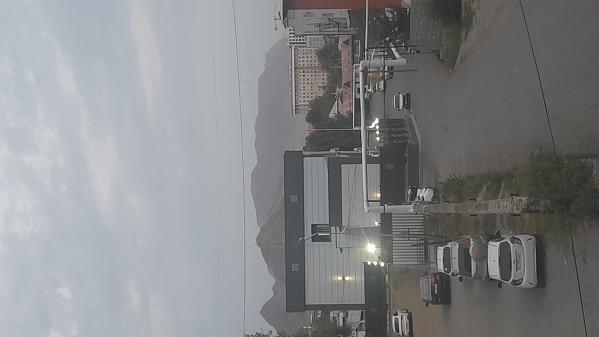 The image shows a street scene in Osh City, Kyrgyzstan. A modern building with a dark facade and white horizontal accents is visible on the left side of the frame. Several cars are parked along the street in front of the building. A white car is parked closest to the foreground. Further down the street, more cars are visible, including a white van driving away from the camera. In the background, a multi-story apartment building with many windows is situated among trees. Mountains loom in the distance, shrouded in haze. The sky is overcast.