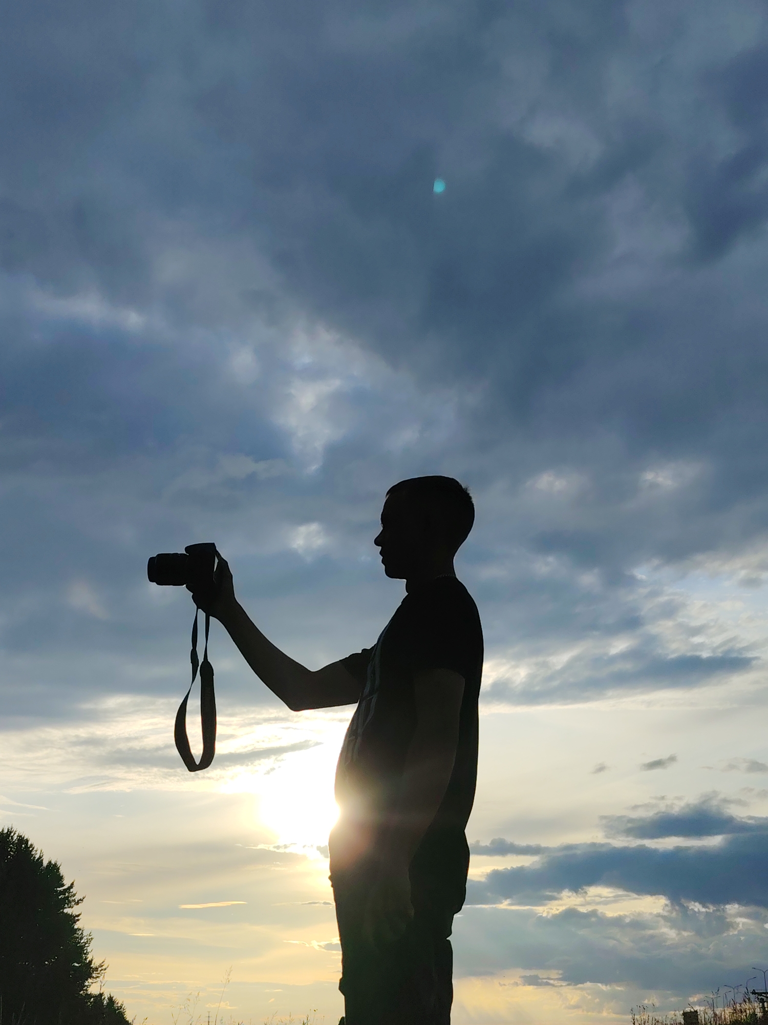 A silhouette of a person holding a camera is captured against a dramatic sky at sunset. The individual, facing to the left, is in profile, with their right arm extended, holding the camera up to eye level as if to take a photograph. A camera strap hangs down from the wrist. The person is wearing a dark t-shirt.
The sky is filled with a mix of dark, brooding clouds and lighter, wispy ones, illuminated by the setting sun. The sun itself is a bright, intense point of light just above the horizon, casting a lens flare that creates a luminous glow. The light filters through the clouds, creating rays and highlighting the texture of the sky.
In the lower left corner, the dark silhouette of trees lines the horizon, suggesting a natural or semi-rural setting. The foreground is also silhouetted, with hints of tall grass or reeds. The overall impression is one of a photographer capturing the beauty of a sunset, possibly in Chaikovsky, Russia. The time of day is clearly sunset, and the weather appears to be partly cloudy, with dramatic light conditions. There is a faint cyan circular artifact visible in the upper portion of the image, which is likely a lens flare or a reflection. No visible text is present in the image.