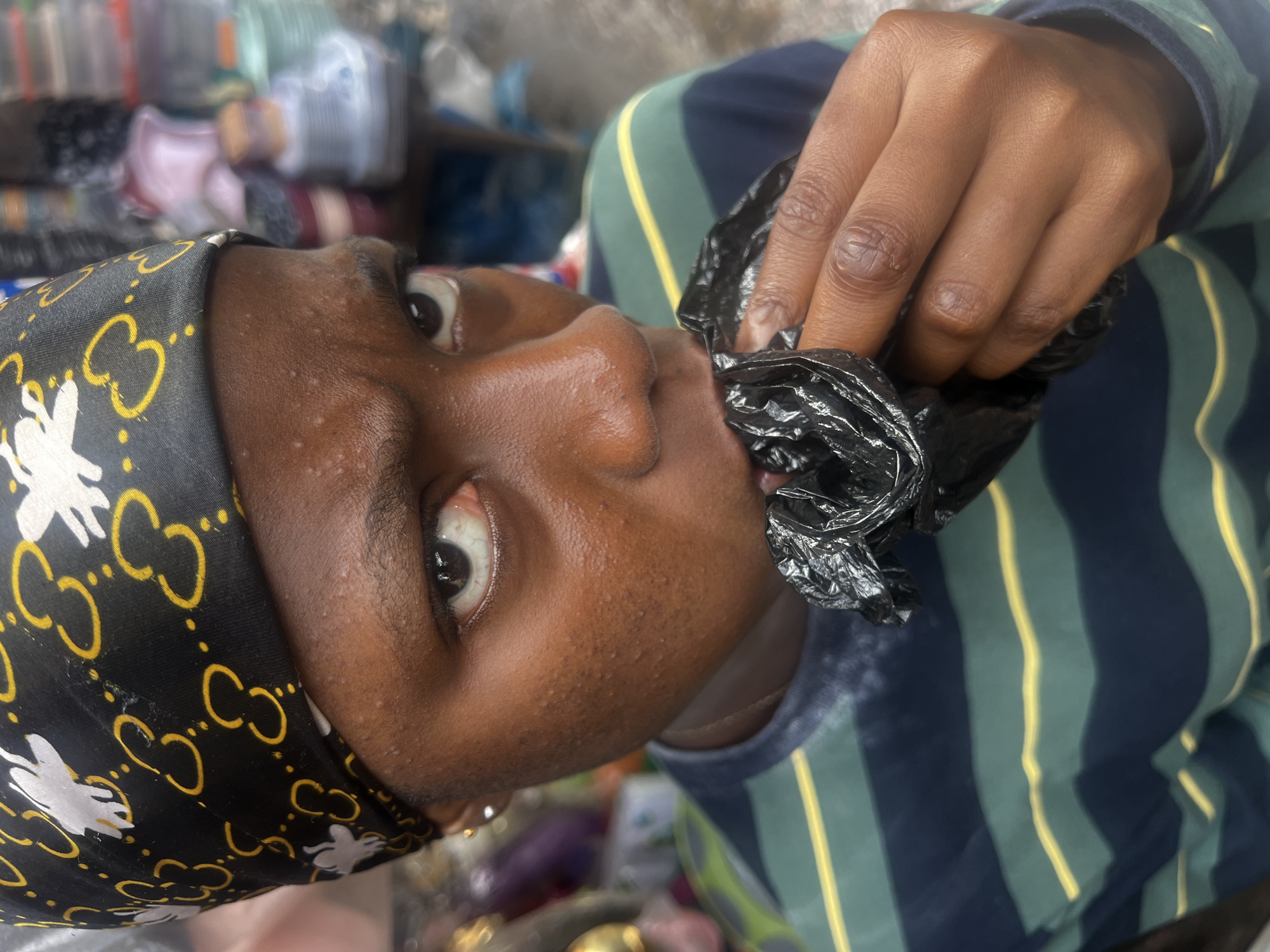 A young person with dark skin wears a black bandana with a yellow and white pattern and a green and blue striped t-shirt. They are holding a crumpled black plastic bag to their mouth, with their right hand to their face. The background is blurred, suggesting a busy market environment. The person's facial expression is solemn, with furrowed brows and their eyes looking directly at the camera. The scene is set in Nkpor, Nigeria.