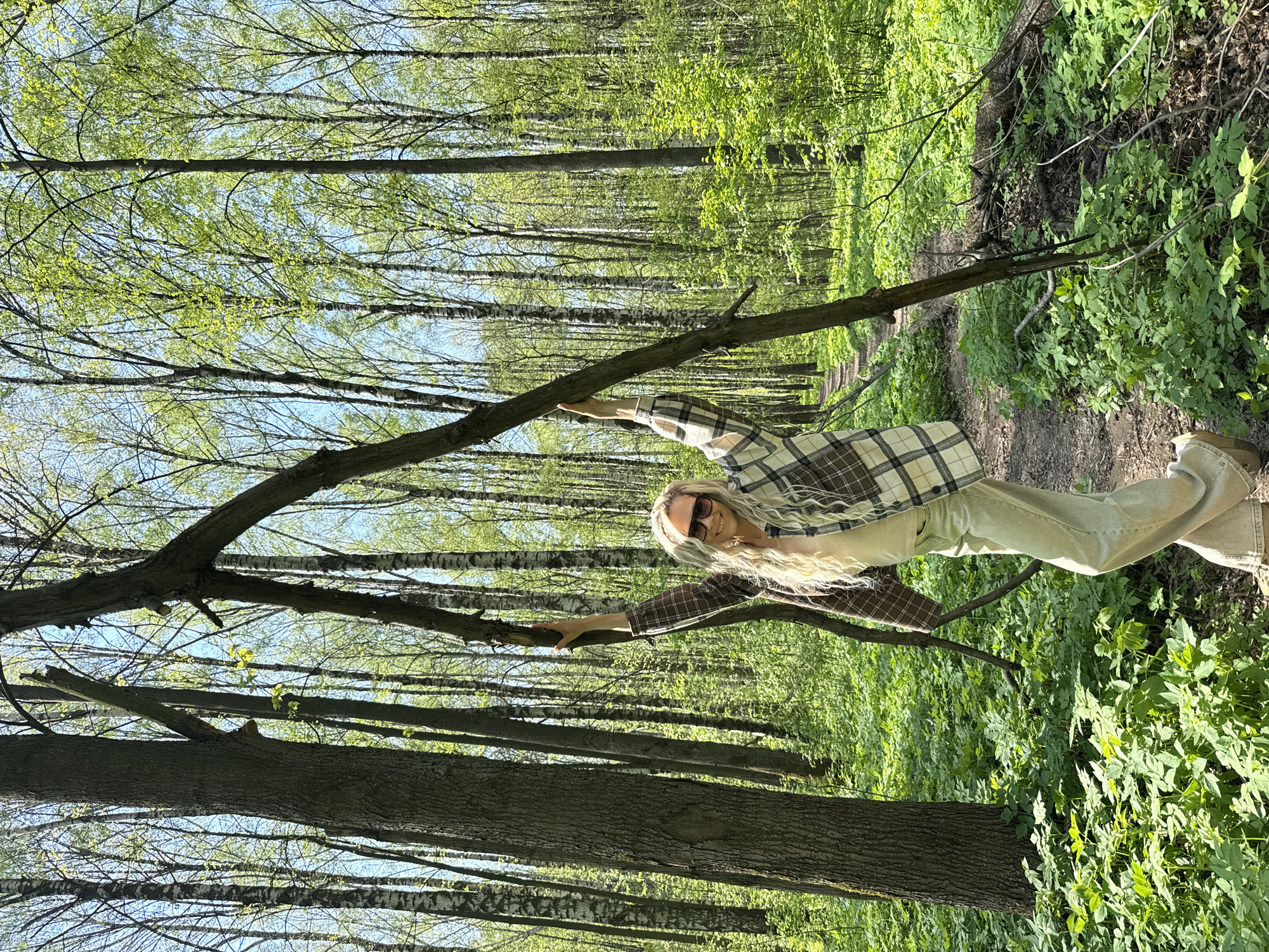 A long-haired blonde woman in sunglasses, a plaid shirt, and light-colored pants stands in a forest. She holds onto a fallen branch with both hands. Sunlight streams through the trees, illuminating green undergrowth and tree trunks. A narrow dirt path can be seen winding through the forest. The location appears to be a wooded area, possibly a park or forest, in Unknown, Unknown.