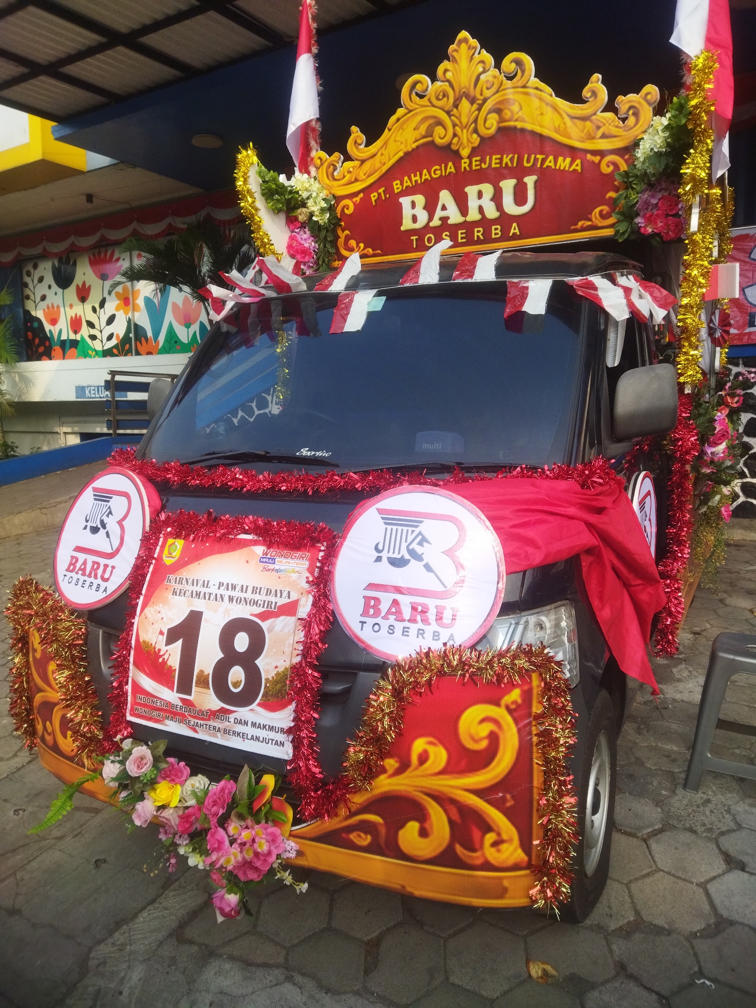 The image shows a black car decorated for a parade in Wonogiri, Indonesia. The car is adorned with red and white Indonesian flags, gold and red tinsel, and flowers. A banner on the front of the car reads "KARNAVAL - PAWAI BUDAYA KECAMATAN WONOGIRI 18". Above the windshield, another banner displays "PT. BAHAGIA REJEKI UTAMA BARU TOSERBA". Several circular logos with the letter "B" and the text "BARU TOSERBA" are attached to the front sides of the car. The car is parked on a paved surface, possibly a street or parking lot, in front of a building with a blue facade and a mural of colorful flowers on the wall. A portion of a gray stool is visible on the right side of the image. The overall scene depicts a festive atmosphere, likely for a cultural event.