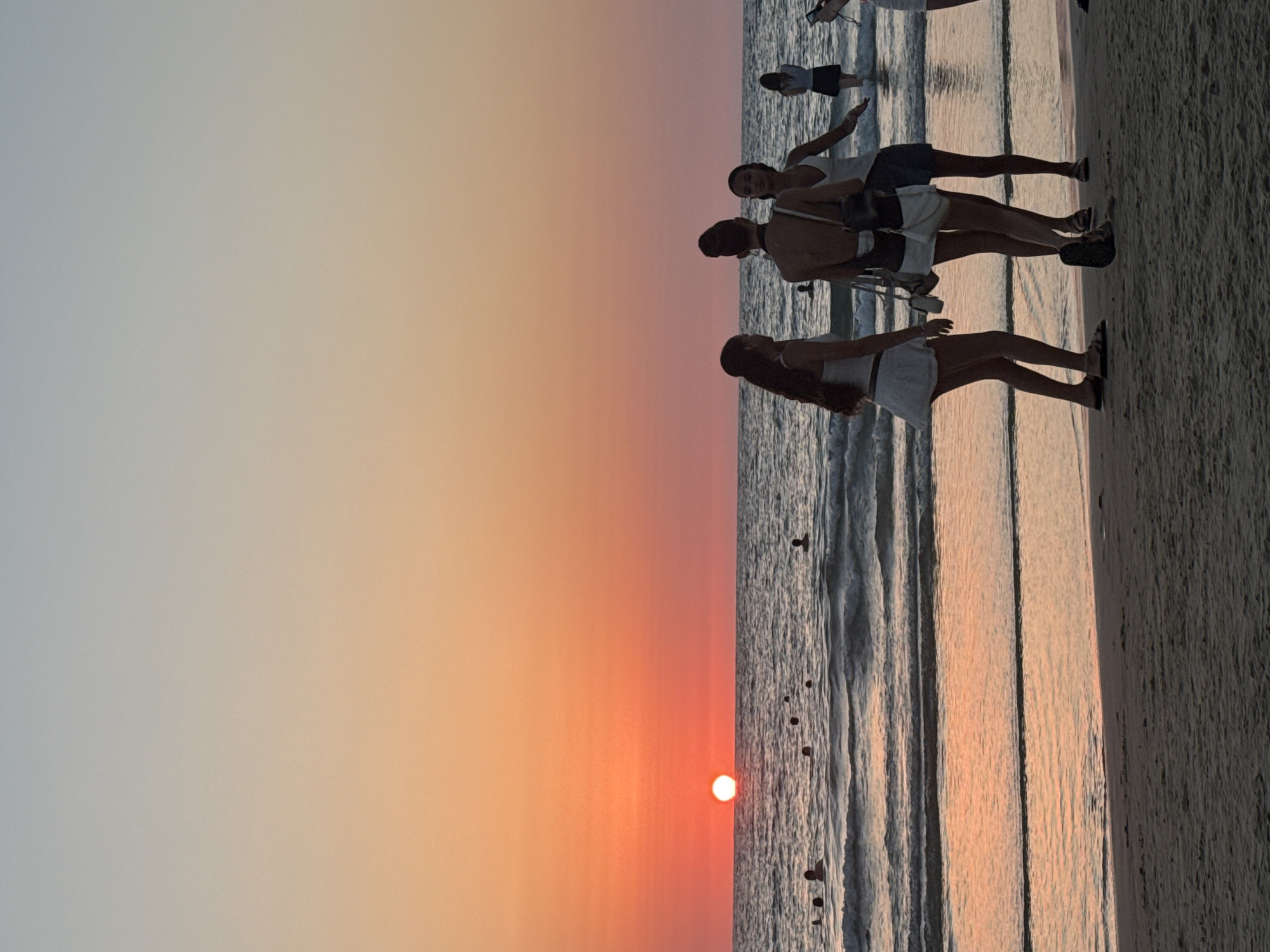 This photograph captures a sunset scene on the beach in Tel Aviv, Israel. The sun, a bright orb, is setting on the horizon, casting an orange and pink glow across the sky and reflecting on the water. Several people are visible in the sea, some swimming and others standing. On the shore, a group of young women are gathered, some facing the sea and others interacting with each other. The sand of the beach is in the foreground, textured and stretching towards the water's edge. The overall atmosphere is serene and idyllic, typical of a summer evening by the Mediterranean Sea.