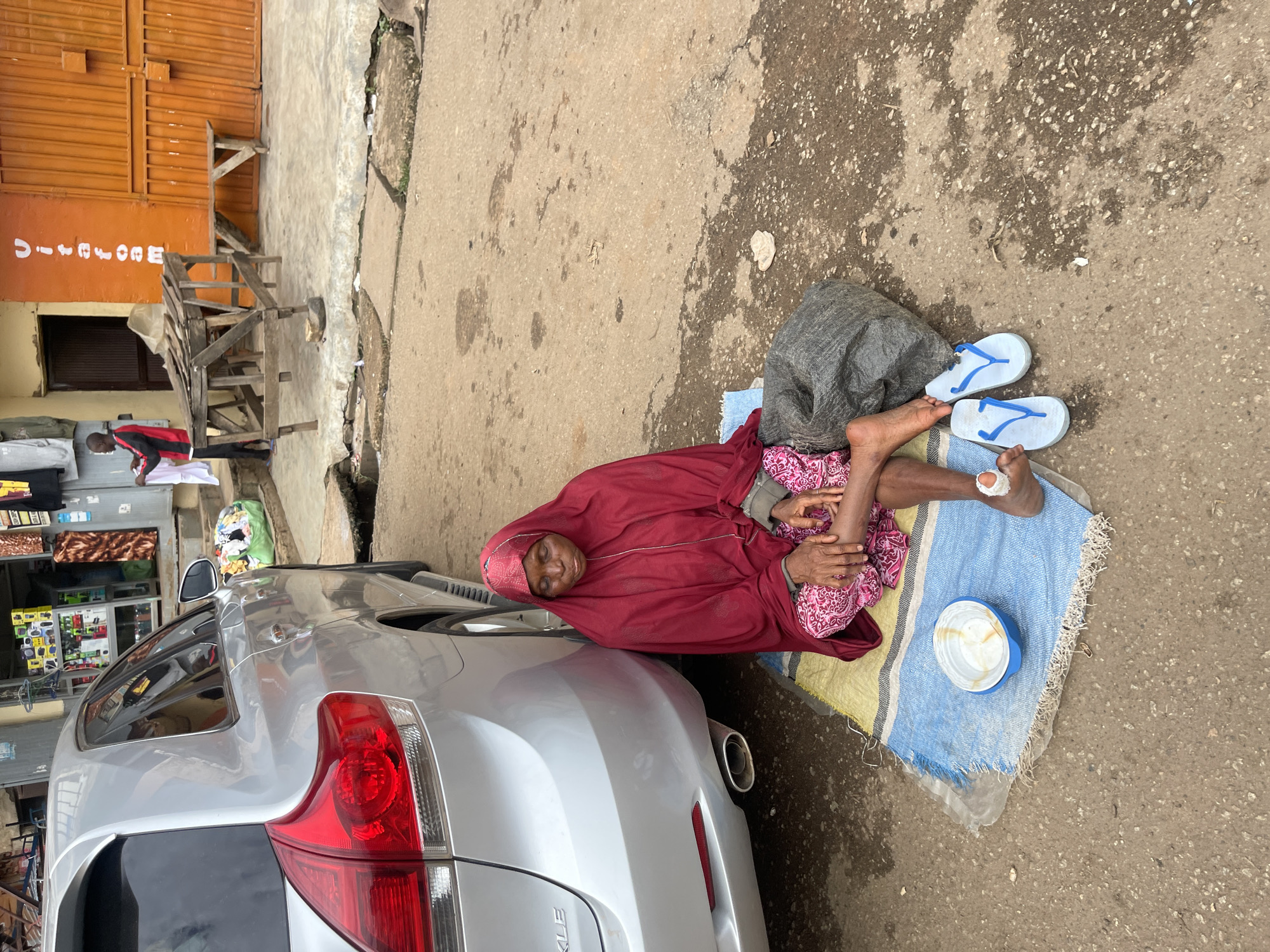 A full shot shows an elderly Nigerian woman sitting on a rug on the ground next to a silver car. She is wearing a red head covering and a pink and white patterned dress. She is looking down at a white bowl on the rug. A large gray bag sits next to her, and a pair of white flip-flops rests on the ground in front of her. Her right foot is bandaged. The scene is set on a street in Jos, Nigeria, with a building behind her that has orange siding and an orange metal gate. A man in a red and black shirt and black pants is standing in front of the building, holding a white garment. A wooden table and bench are set up in front of the building.