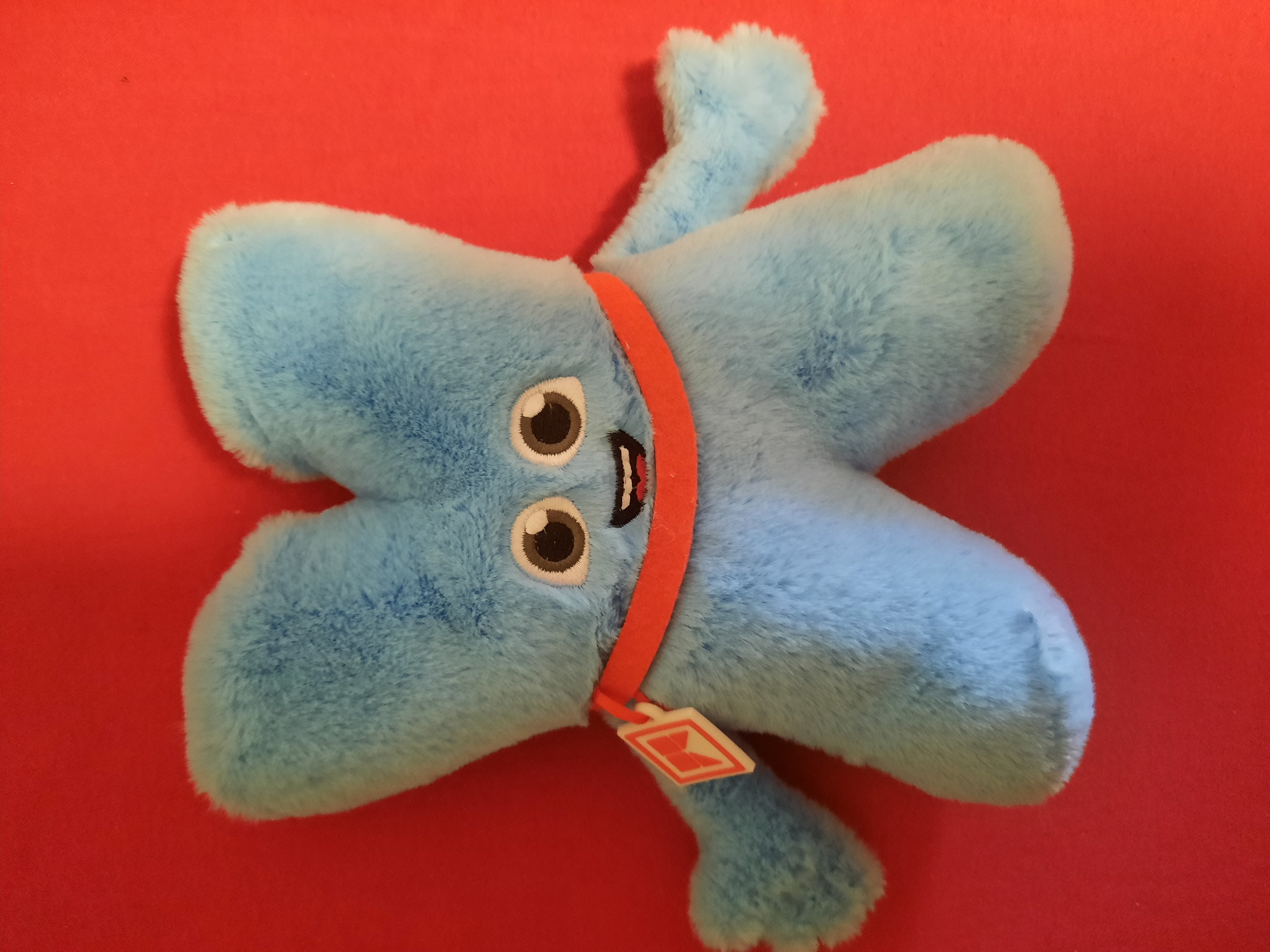 A light blue, plush toy character resembling the letter X is positioned centrally on a vibrant red background. The toy has two large, round eyes with black pupils and white highlights, and a wide, smiling mouth. It wears an orange band around its neck, and a white tag with a red diamond symbol hangs from the left side of the band. The toy's limbs are short and rounded. The background is a flat, solid red surface. The image does not contain any identifiable landmarks or features that would place it in Rosenheim, Germany.