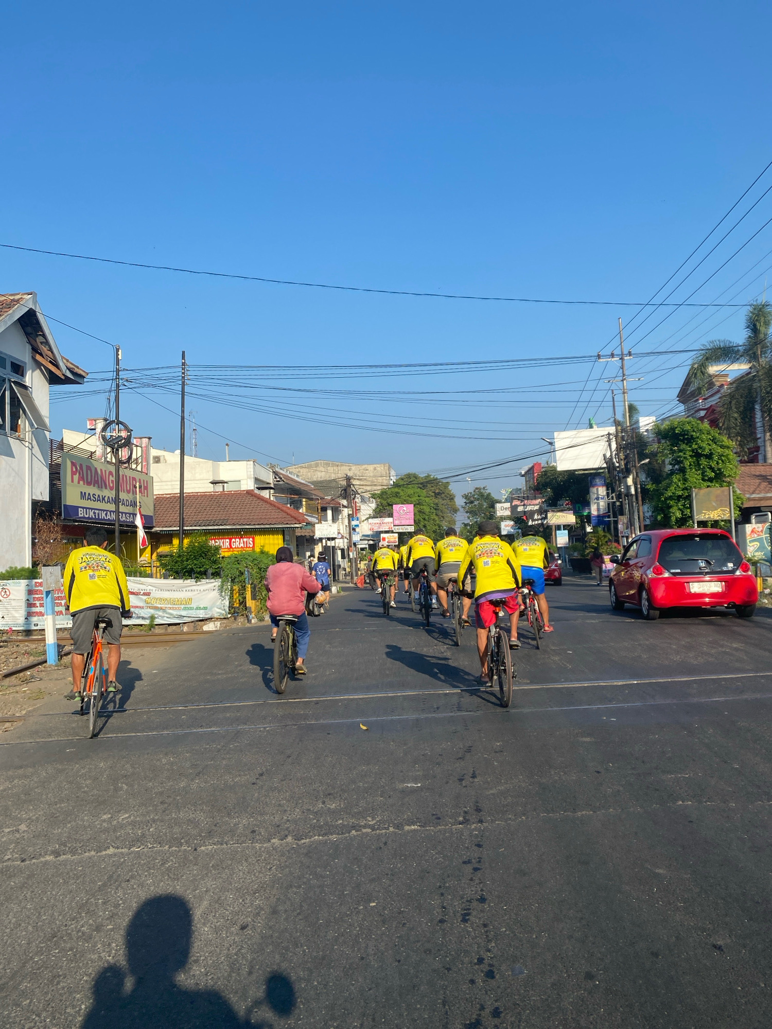 This image captures a group of cyclists wearing bright yellow shirts on a city street in Kediri, Indonesia. The cyclists are moving away from the camera, with the majority in the foreground and middle ground. One cyclist in the far left foreground is wearing a yellow shirt and grey shorts, riding an orange bicycle. Behind him, another cyclist in a pink top and dark pants is riding a bicycle. Further down the street, a procession of cyclists in yellow shirts are visible, some with blue shorts and others with red shorts.
The environment is urban, with buildings lining both sides of the street. A prominent yellow and blue sign for "PADANG MURAH" (cheap Padang cuisine) is visible on the left. There are utility poles and wires overhead, characteristic of an Indonesian streetscape. A red car is seen on the right side of the road, moving in the same direction as the cyclists. The road is asphalt with visible lane markings and some debris. The sky is clear and blue, indicating a sunny day. The overall scene suggests a recreational cycling event or a group commute through the city.