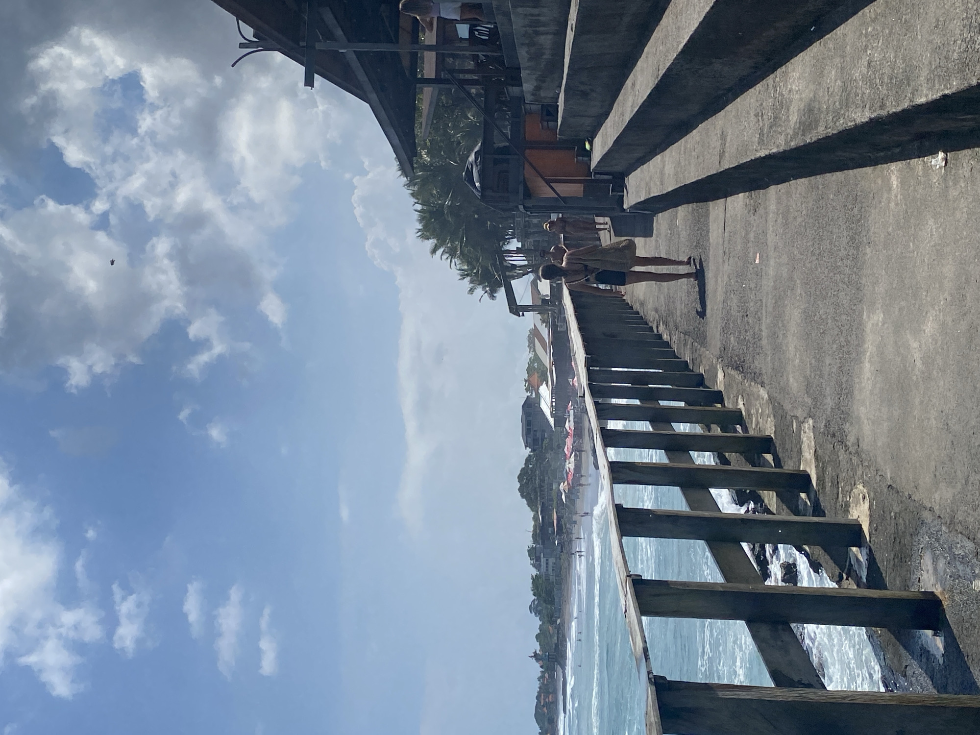 The image shows a beach scene in North Kuta, Indonesia. A concrete pier juts out into the ocean. Steps lead down from the pier into the water. The wooden steps are partially submerged. A row of palm trees is visible in the background. People stand on the pier, looking at the beach. A concrete structure with steps runs parallel to the pier, casting shadows. The sky is blue with scattered white clouds. A small bird is visible in the sky. The beach is lined with buildings and trees. Waves are breaking on the shore.