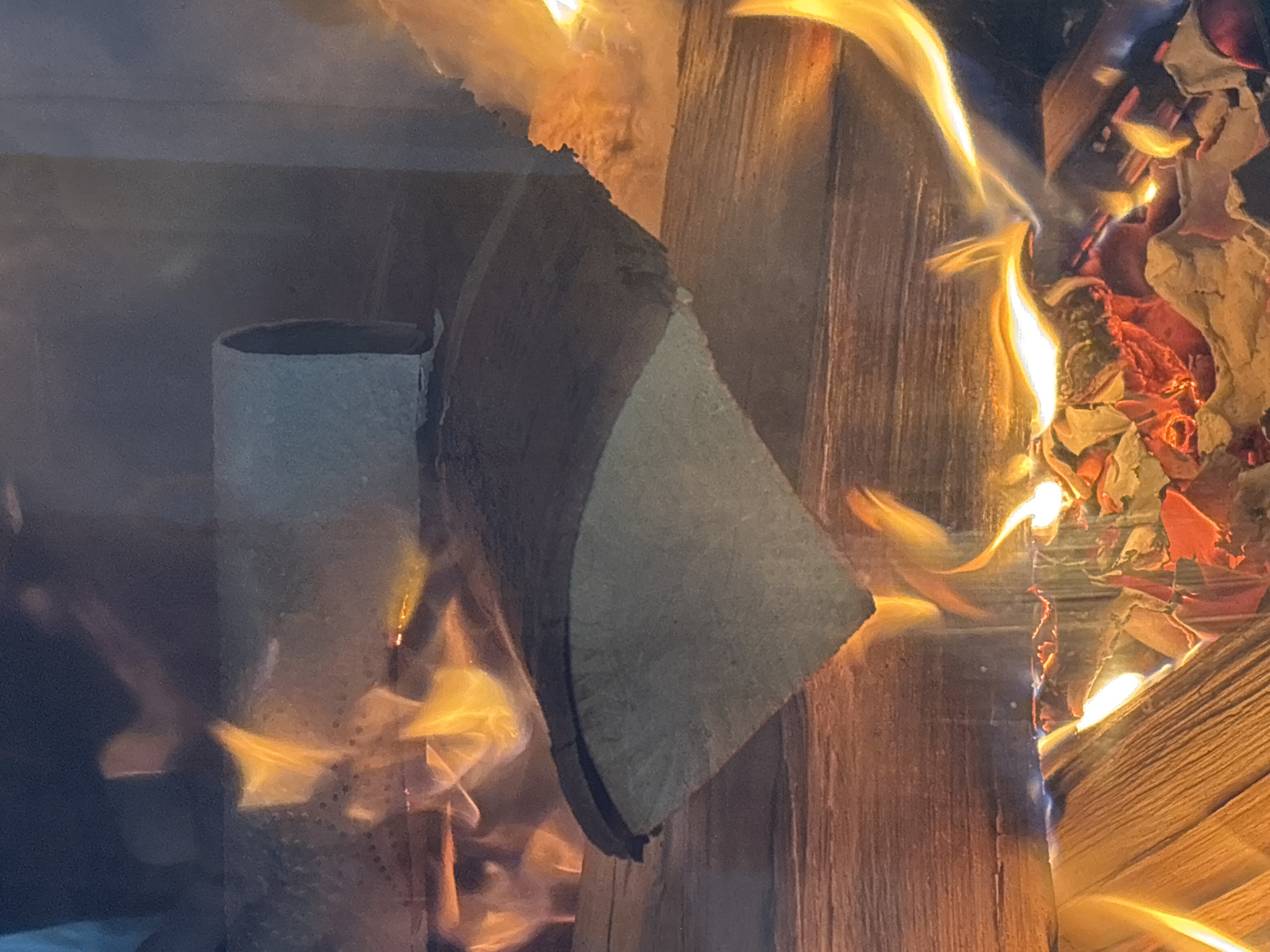 The image shows a close-up view of a fire burning inside a fireplace. The fire is intense, with flames licking upwards and illuminating the surrounding wooden logs. One log, partially burned, rests on top of other logs. The background is dark, suggesting the interior of a room. The scene depicts a cozy and warm atmosphere, typical of a fireplace in a home. There are no people visible in the image. The context of Zvenigorod, Russia, is not directly inferable from the visual elements of the photograph.