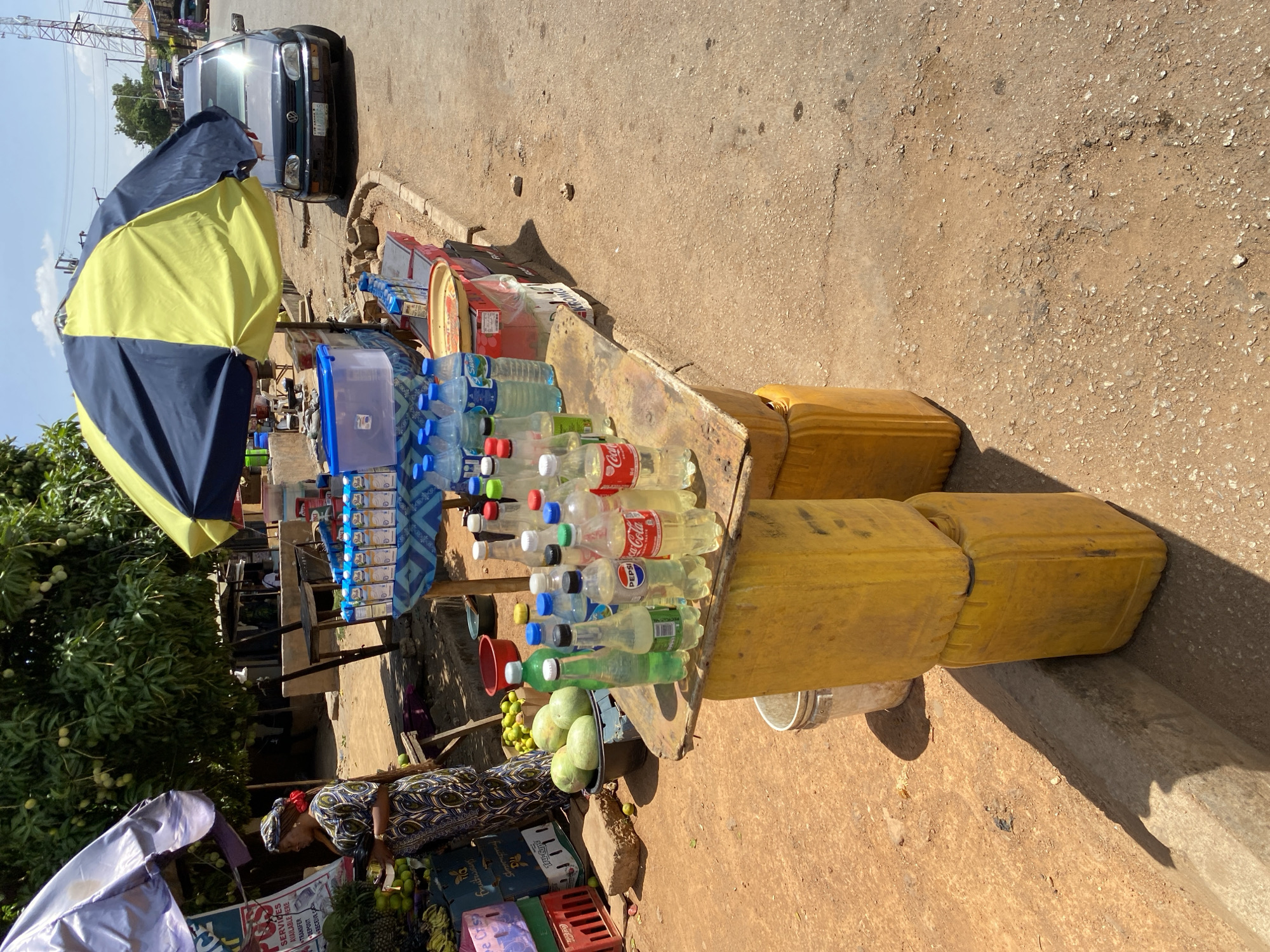 The image depicts a roadside vending setup in Bukuru, Nigeria. The vendor, an African woman wearing a patterned dress and headwrap, stands behind a makeshift table propped up by stacked yellow jerrycans. The table displays various bottled drinks, including water, Coca-Cola, and Pepsi. A blue and yellow umbrella offers shade.
To the left, more produce is visible, suggesting fruits and vegetables are also for sale. The image captures the vendor interacting with the produce stand. A car is parked nearby. The surface under the table is a combination of dirt and concrete. The OCR identifies commercial elements such as "SERVICES AVAILABLE HERE," "TRANSFER," "DEPOSIT," "RECEIVE CASH," and various brand names like "Coca-Cola" and "PEPSI."