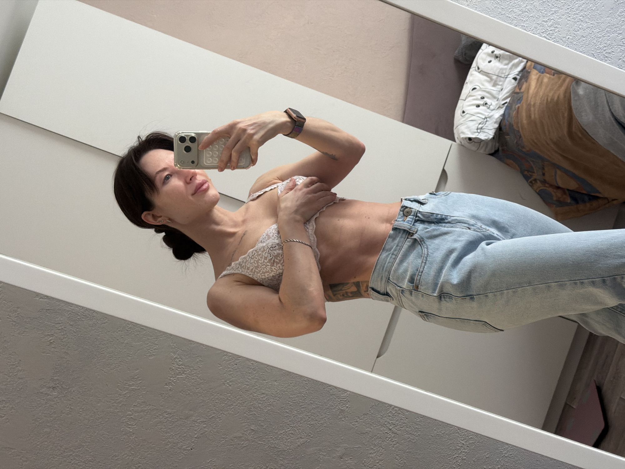 A woman is taking a selfie in front of a mirror. She is wearing jeans and a lace bra. She is holding a phone in her right hand. She has a watch on her left wrist. The background includes a white dresser or wardrobe and a wall with textured paint. Based on visual data, the location is identified as Perm, Russia.