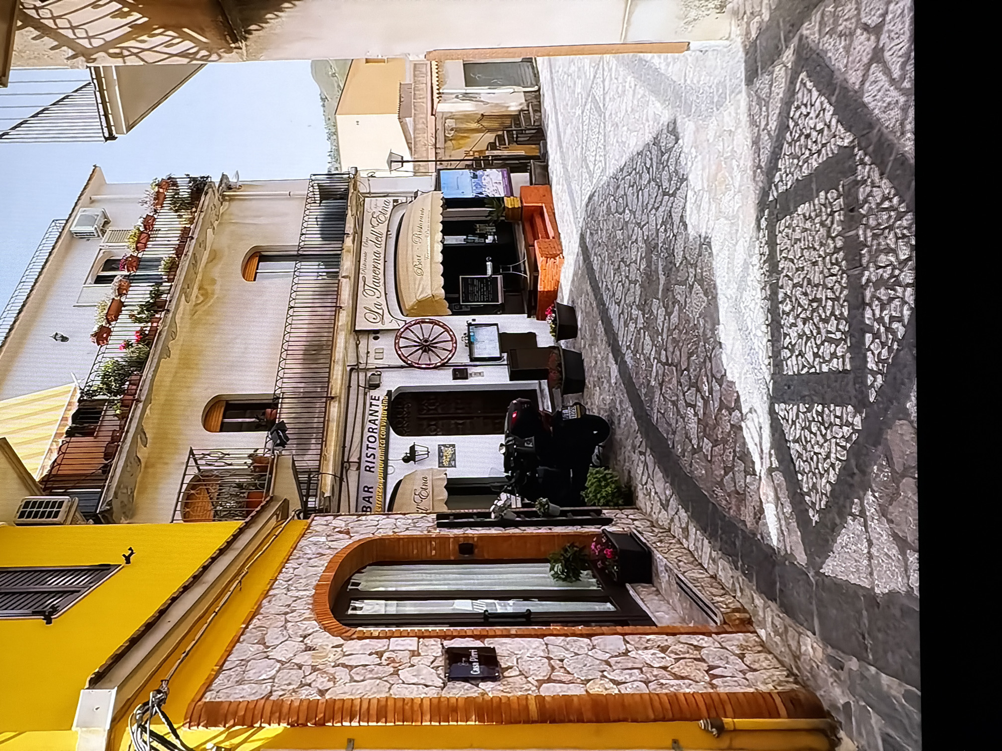 The image depicts a quaint street scene in a Mediterranean-style town, likely in Italy, characterized by its architecture and cobblestone pathways. The focus is on the facade of a building with a prominent yellow section on the left, adorned with a stone-clad lower level and an arched doorway. To the right, a building painted in a light, possibly off-white, color features balconies overflowing with potted plants, an air conditioning unit, and a banner above a restaurant entrance that reads "La Taverna dell'Etna". A black scooter is parked near the restaurant, and the street itself is paved with irregular stones, forming a textured pattern. Sunlight casts shadows across the pavement, suggesting it is daytime. The overall impression is one of a charming, sun-drenched European village street. There is no indication that this scene is located in Chirchik, Uzbekistan.