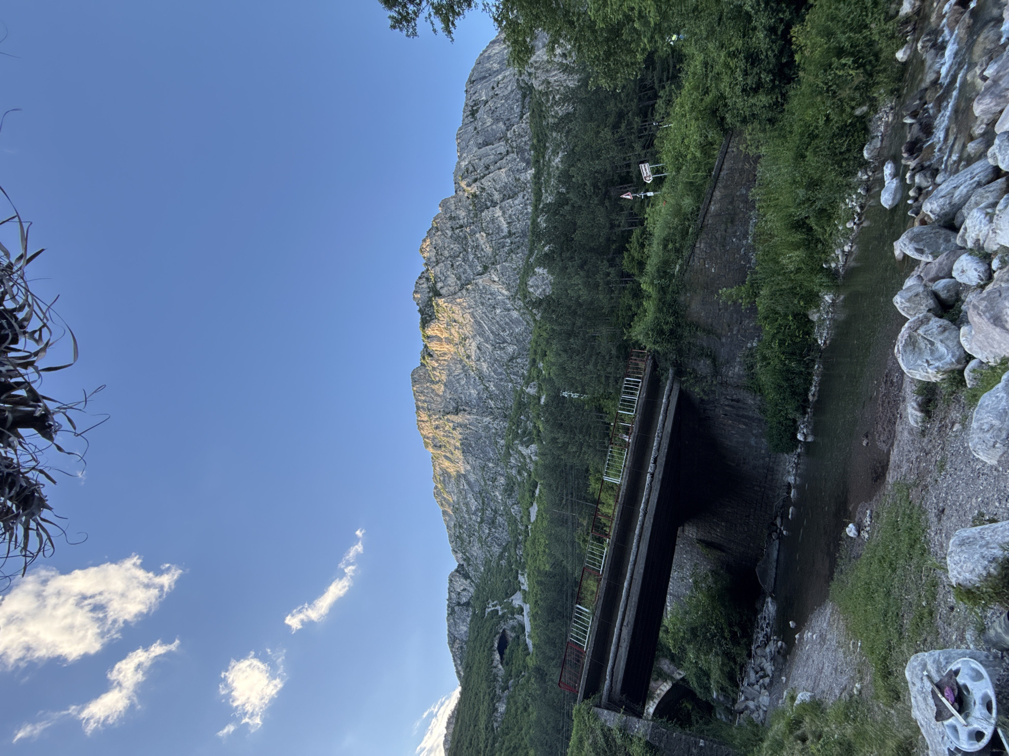 The image captures a scenic view of a mountain range and a bridge over a river, with vegetation covering the slopes. The mountain's rocky facade is illuminated by sunlight, creating a dramatic contrast with the shadows. Trees and bushes are abundant, particularly on the lower slopes and along the riverbanks. A concrete bridge, featuring red and white railings, spans the river, which flows over a bed of stones. The sky is a clear blue with a few scattered clouds. The scene suggests a natural, possibly remote location, with the presence of a bridge indicating a pathway or road access.