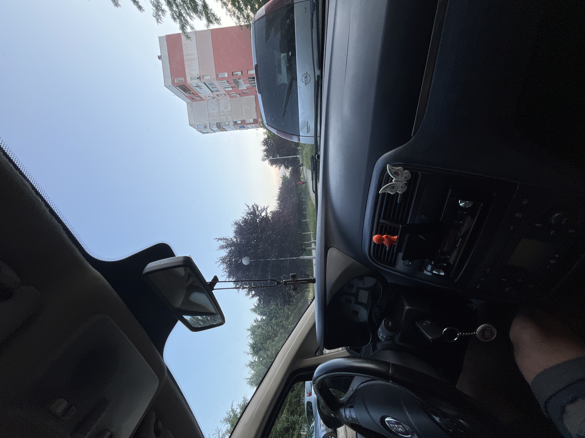 The image captures a view from inside a stationary car, looking out through the front windshield towards an urban or suburban residential area in Plovdiv, Bulgaria. The lighting is soft and diffused, suggesting either early morning or late afternoon.
To the left of the frame, a multi-story apartment building with alternating sections of pinkish-red and beige/grey facade is visible against a clear, bright blue sky. Directly ahead, a silver Opel Zafira car is parked or stationary, with its "Opel" logo and "ZAFIRA" text clearly visible on the rear. Beyond the Opel, a line of green trees, some with reddish foliage, and a street light are present, hinting at a street or park area.
The interior of the observer's car occupies the right and lower portions of the image. A black dashboard with various controls and a central console is prominent. A silver butterfly-shaped air freshener clip is attached to an air vent on the far right of the dashboard, and a small orange figure is clipped near the central console. The steering wheel, featuring a "FIAT" logo, is visible in the lower right, along with a partial view of a leg. A set of car keys, attached to a "FIAT" branded keychain, hangs from the ignition. The rearview mirror is positioned at the top center of the windshield, reflecting parts of the car's ceiling.
No people are distinctly visible, and the scene presents a calm, observational mood of an everyday urban setting.